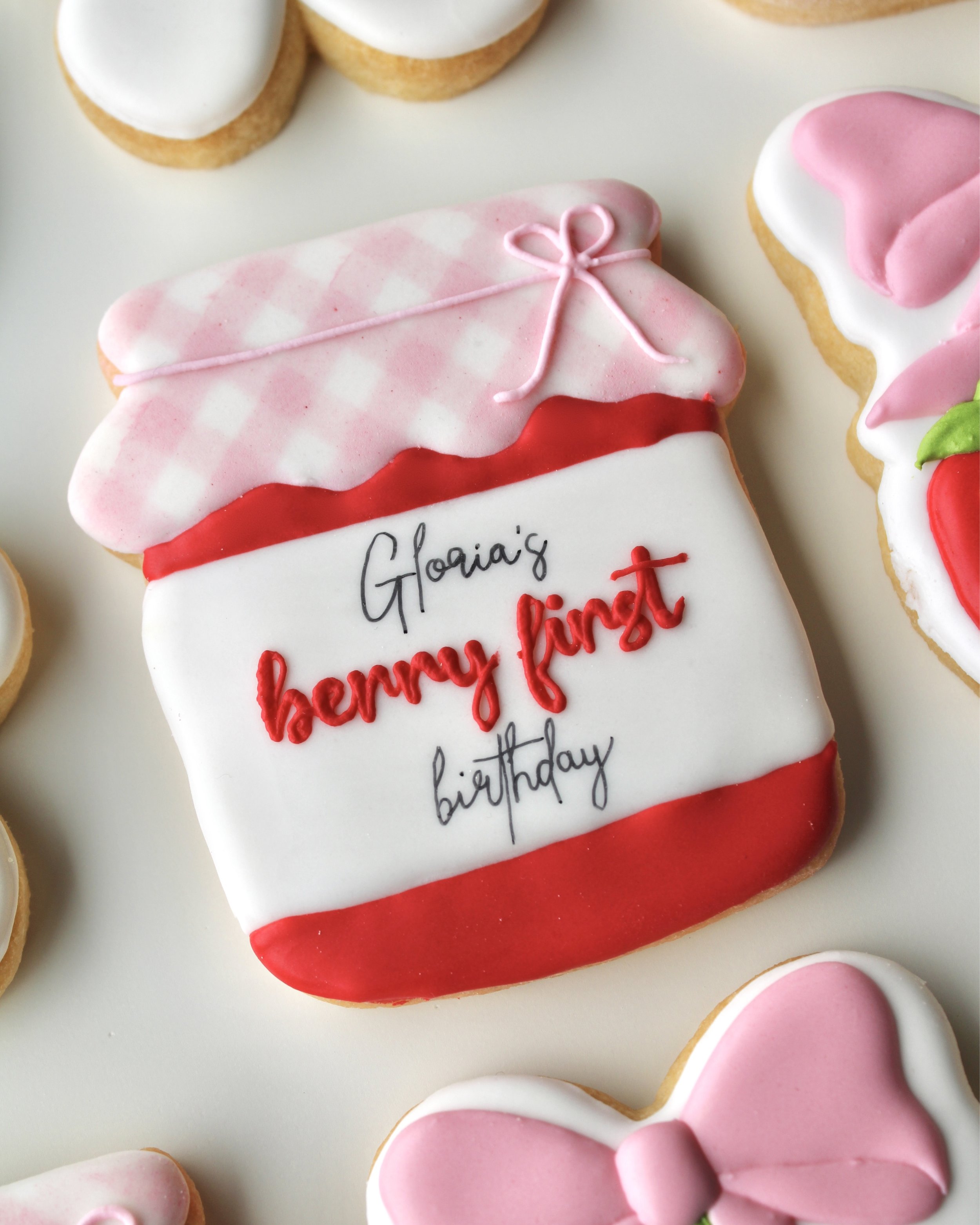 Close-up of strawberry jam jar cookie with red icing, pink and white gingham print, and “Gloria’s Berry First Birthday” written on the label.