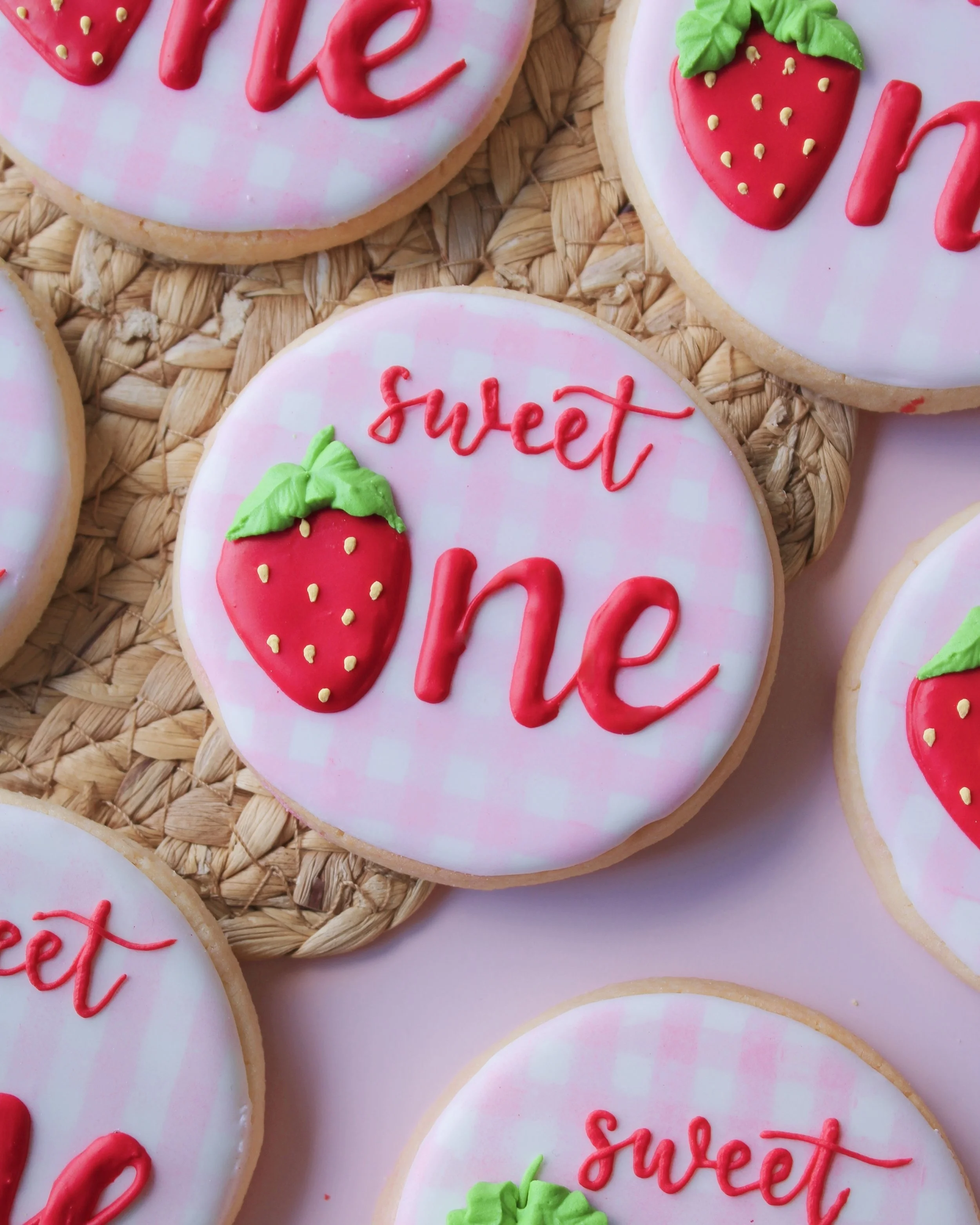 Close-up of “Sweet One” themed cookie with pink and white gingham icing and “one” written in red icing, featuring a strawberry-shaped “o”.