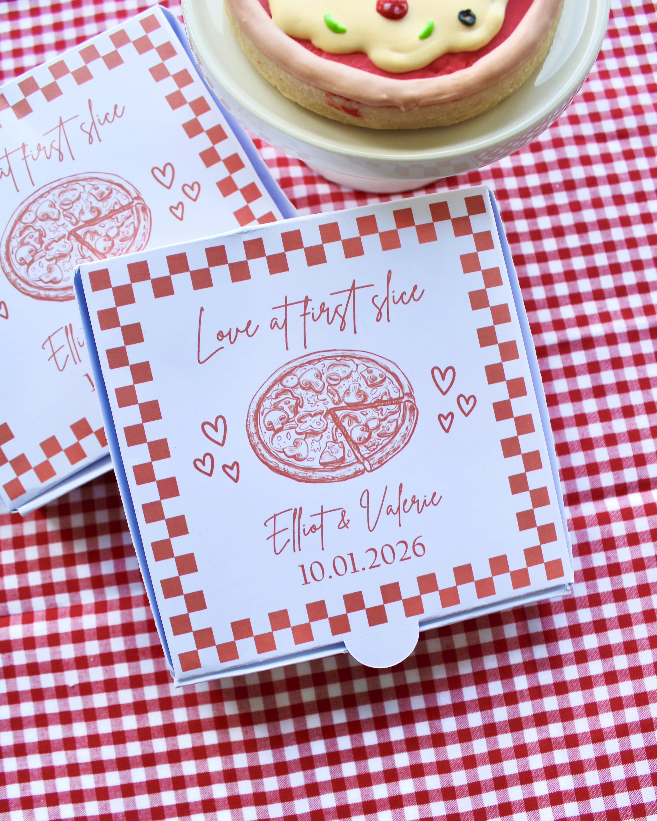 Close-up of a mini pizza box reading “Love at First Slice” with a pizza illustration on the lid, styled on red and white checkered fabric.
