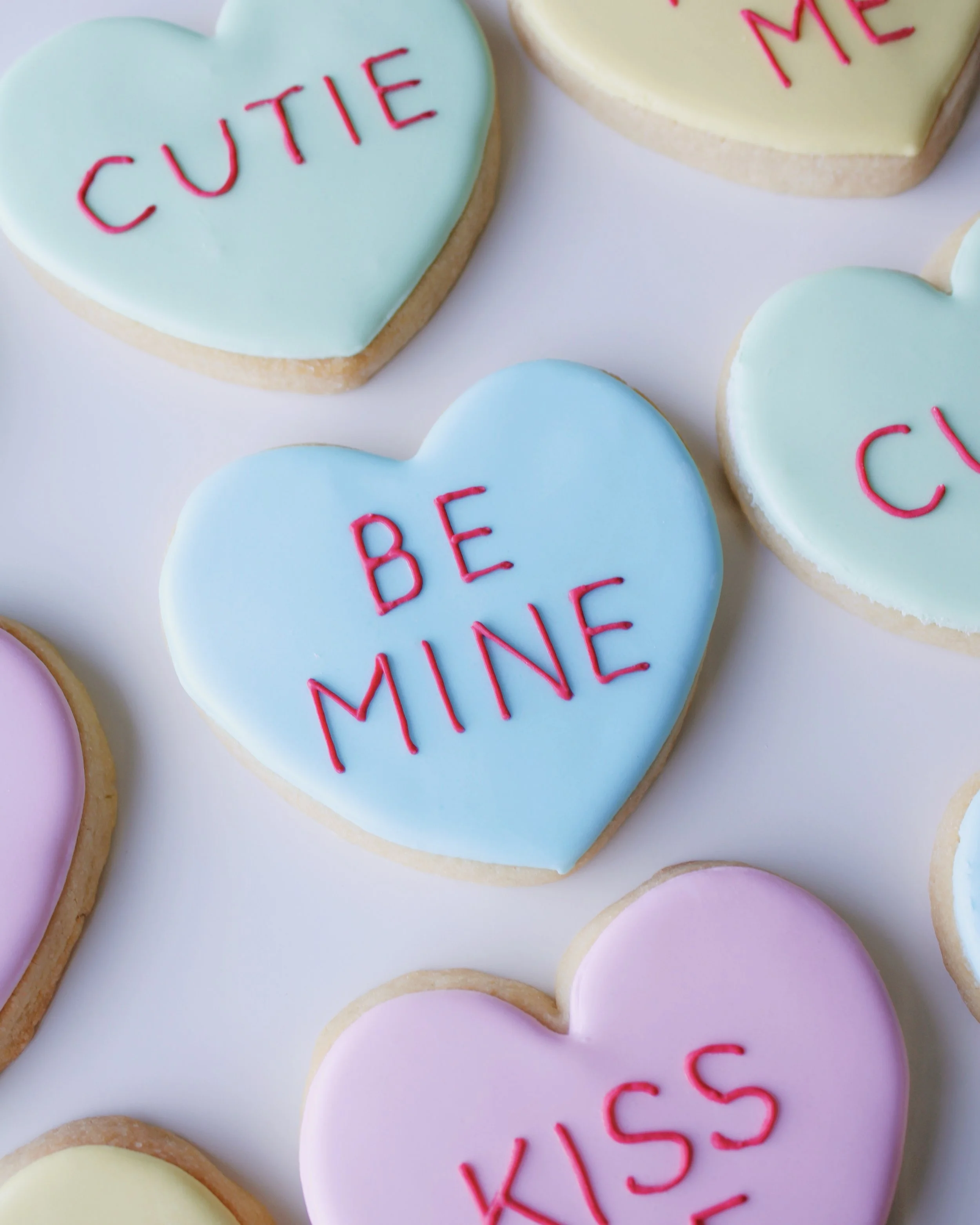 Close-up of a heart-shaped cookie decorated like a conversation heart candy in pastel blue icing with red text reading “be mine.”