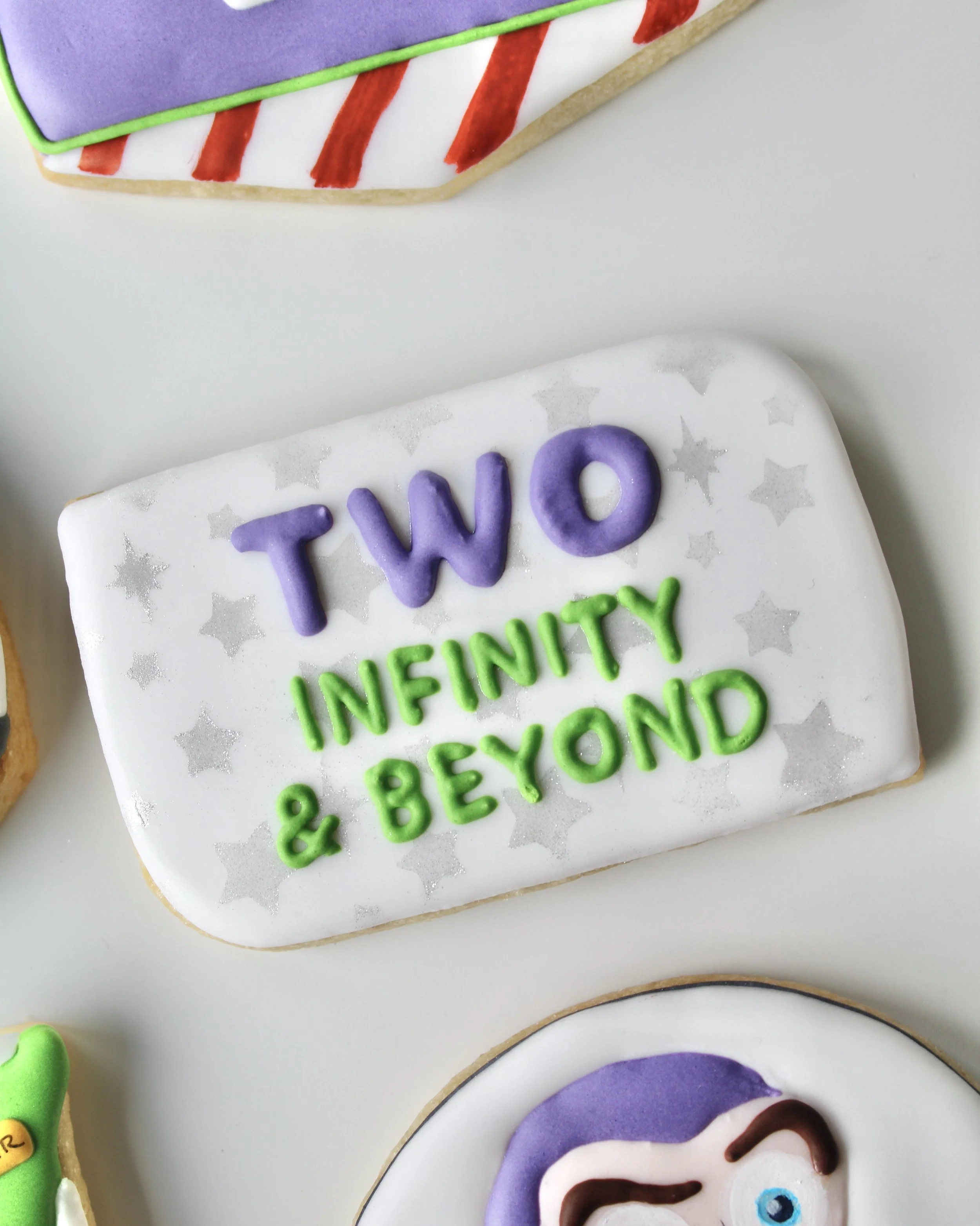 Close-up of a white cookie decorated with silver stars and “Two infinity & beyond” in purple and green royal icing.