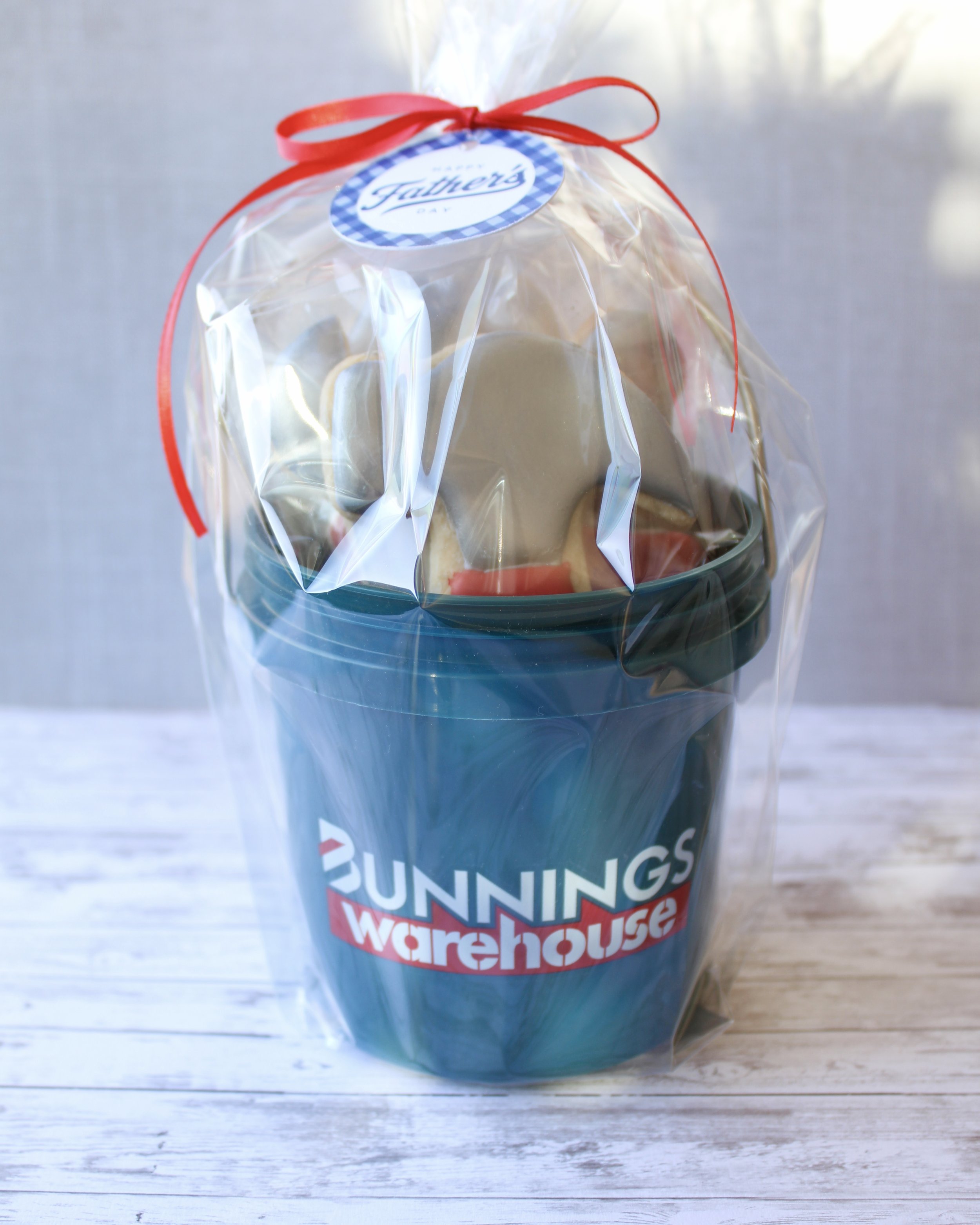 Father's Day tool cookies in a dark green Bunnings Warehouse mini bucket, wrapped in cellophane with a red ribbon and Happy Father's Day gift tag on a grey washed wooden background.