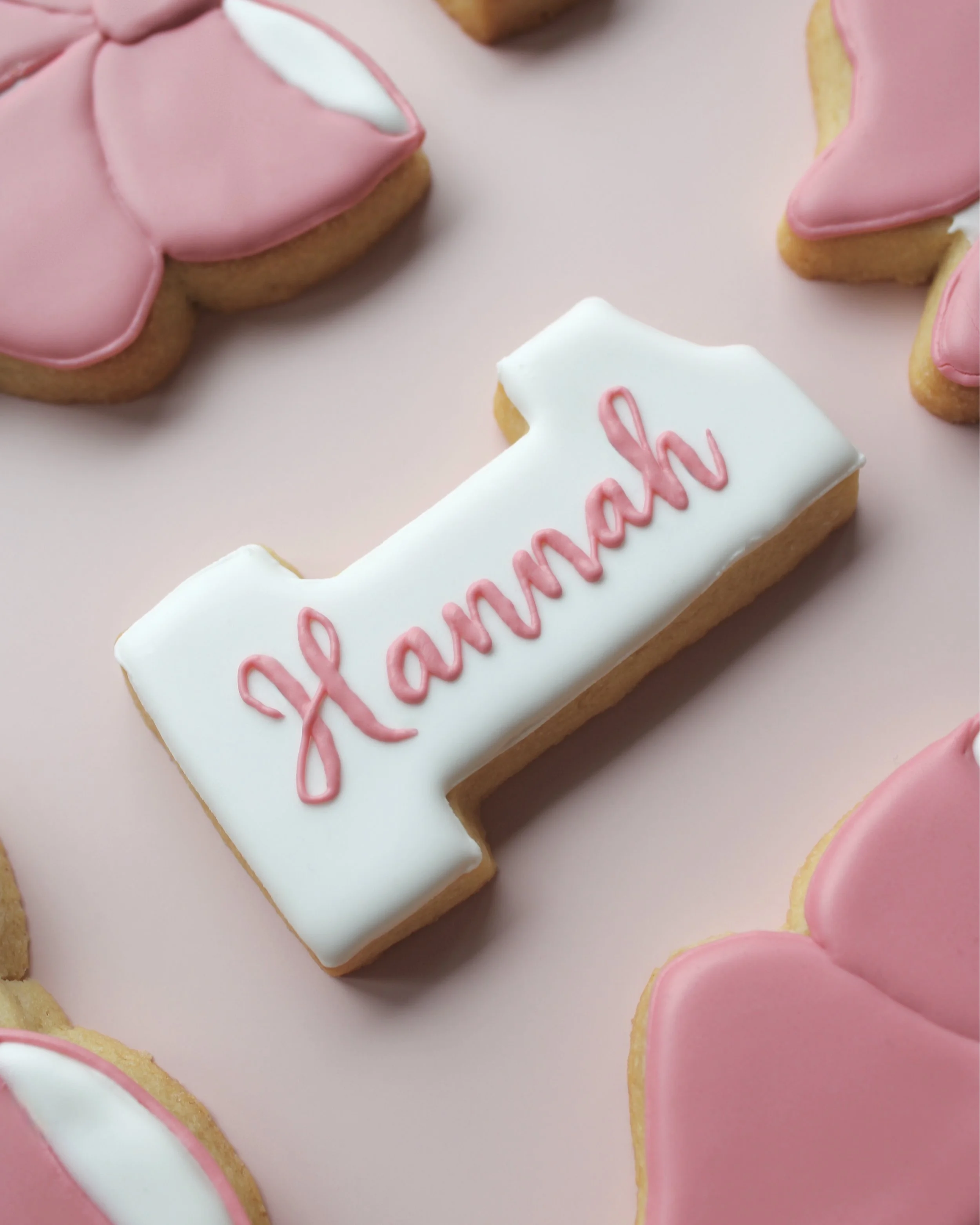 Close-up of white number 1 cookie with “Hannah” written in blush script.