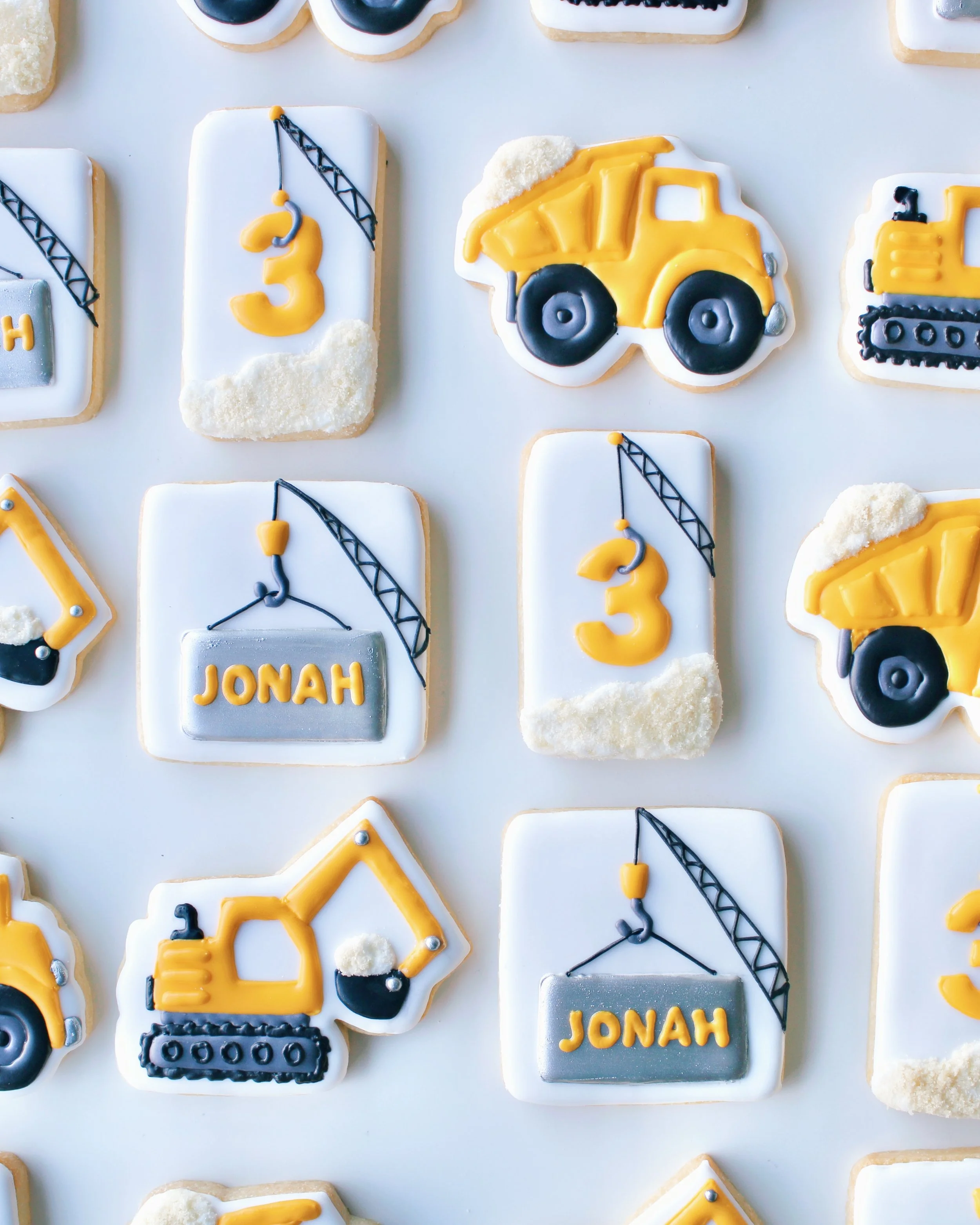 Construction Birthday Cookie Set featuring trucks, excavators and cranes in golden yellow, white, silver and black with personalised name and age cookies.
