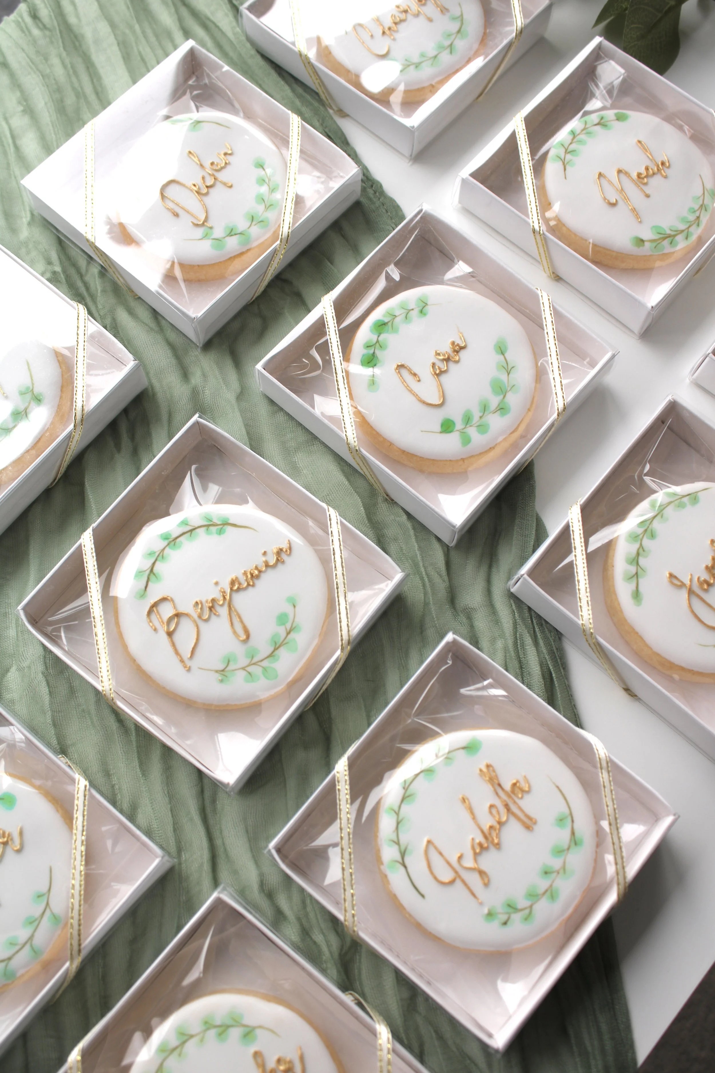 Personalised eucalyptus wedding place card cookies laid out on a styled background featuring hand-painted watercolour greenery and edible gold names