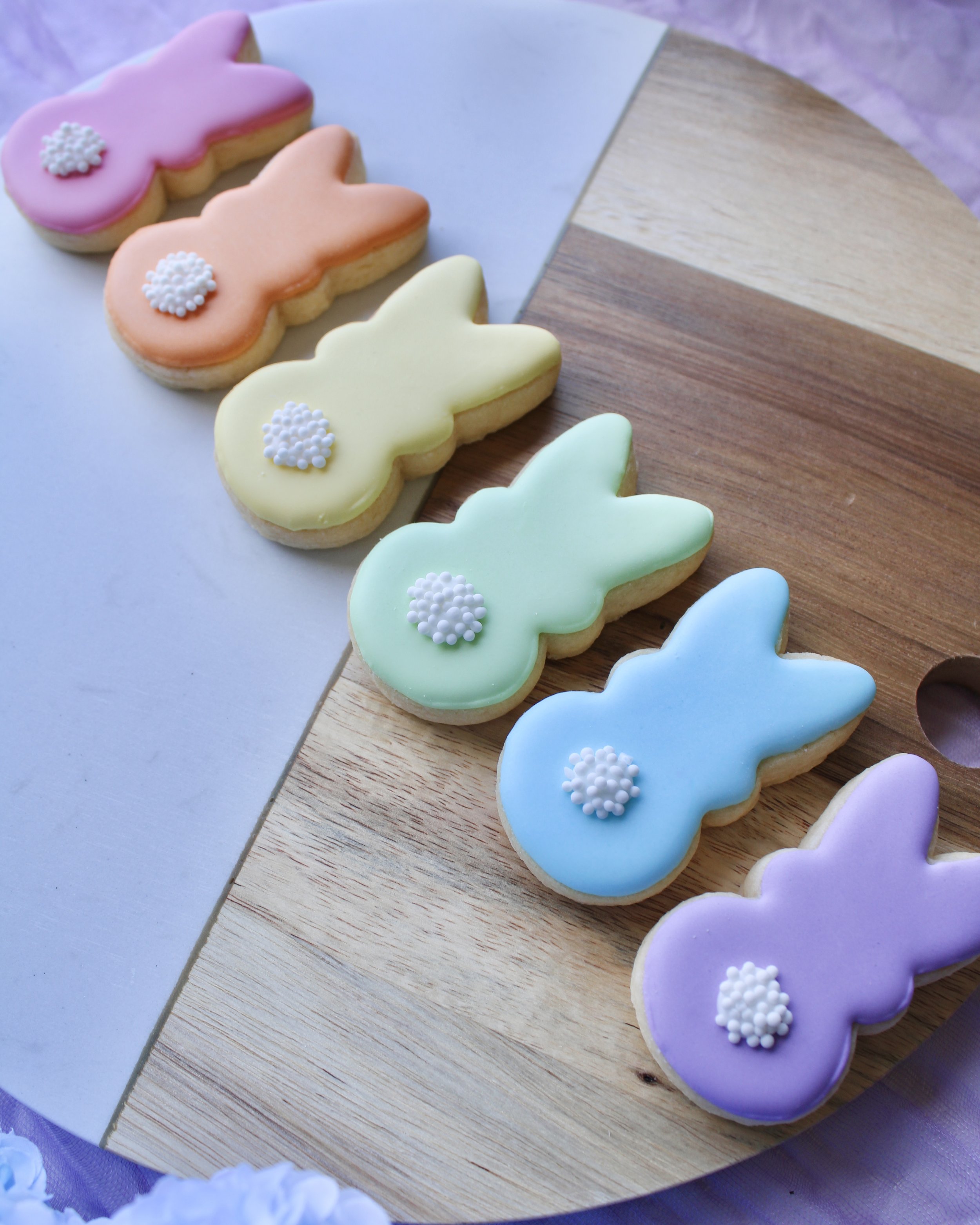 Six mini Easter Bunny Peeps cookies in rainbow pastel colours with white tails on a marble and wood board, styled with lilac fabric, Easter eggs, hydrangeas and a chalkboard sign.