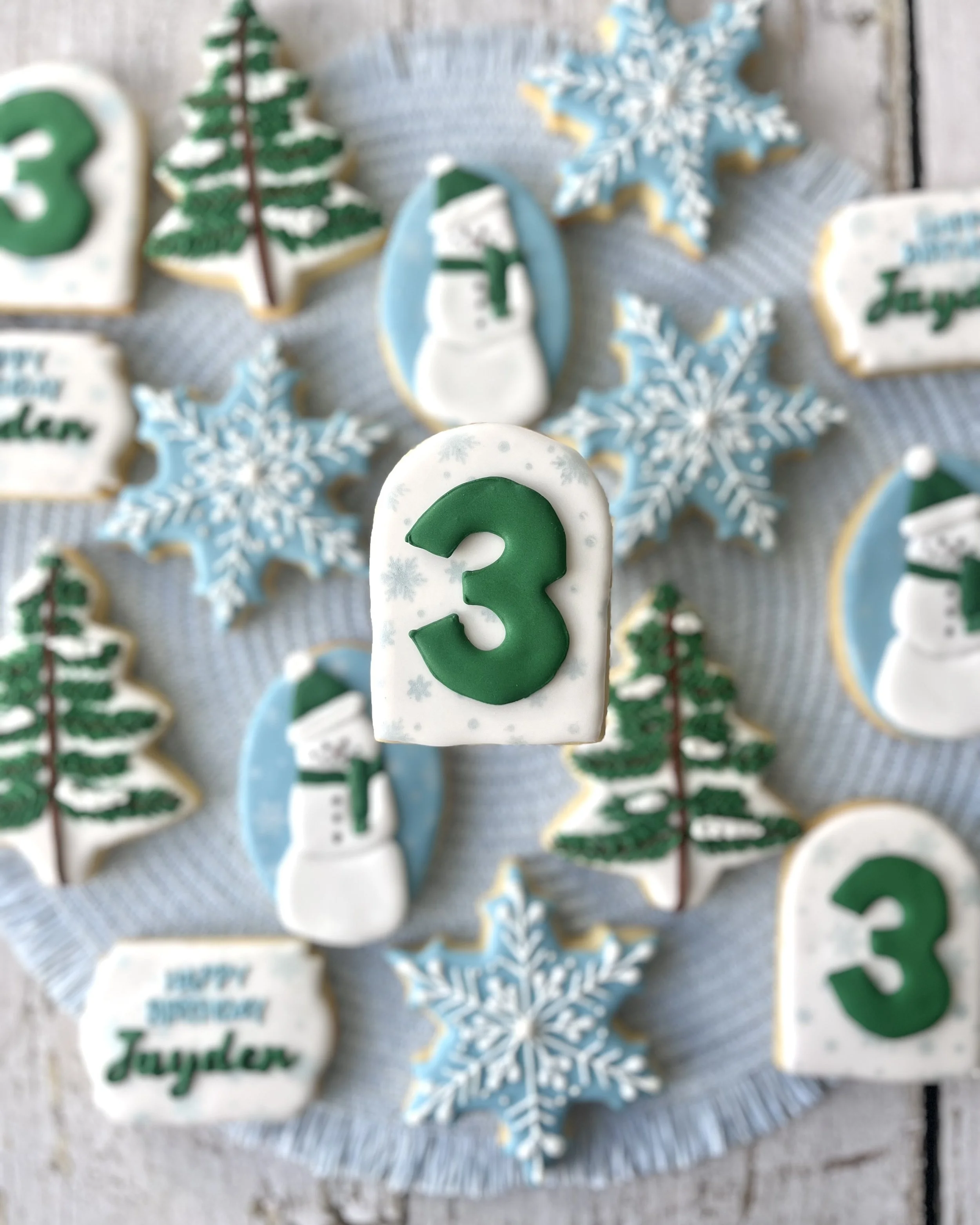 Winter Wonderland 3rd birthday cookie set featuring blues, deep greens and whites with snowflakes, snowman, “Happy Birthday Jayden,” number 3 and pine tree designs.