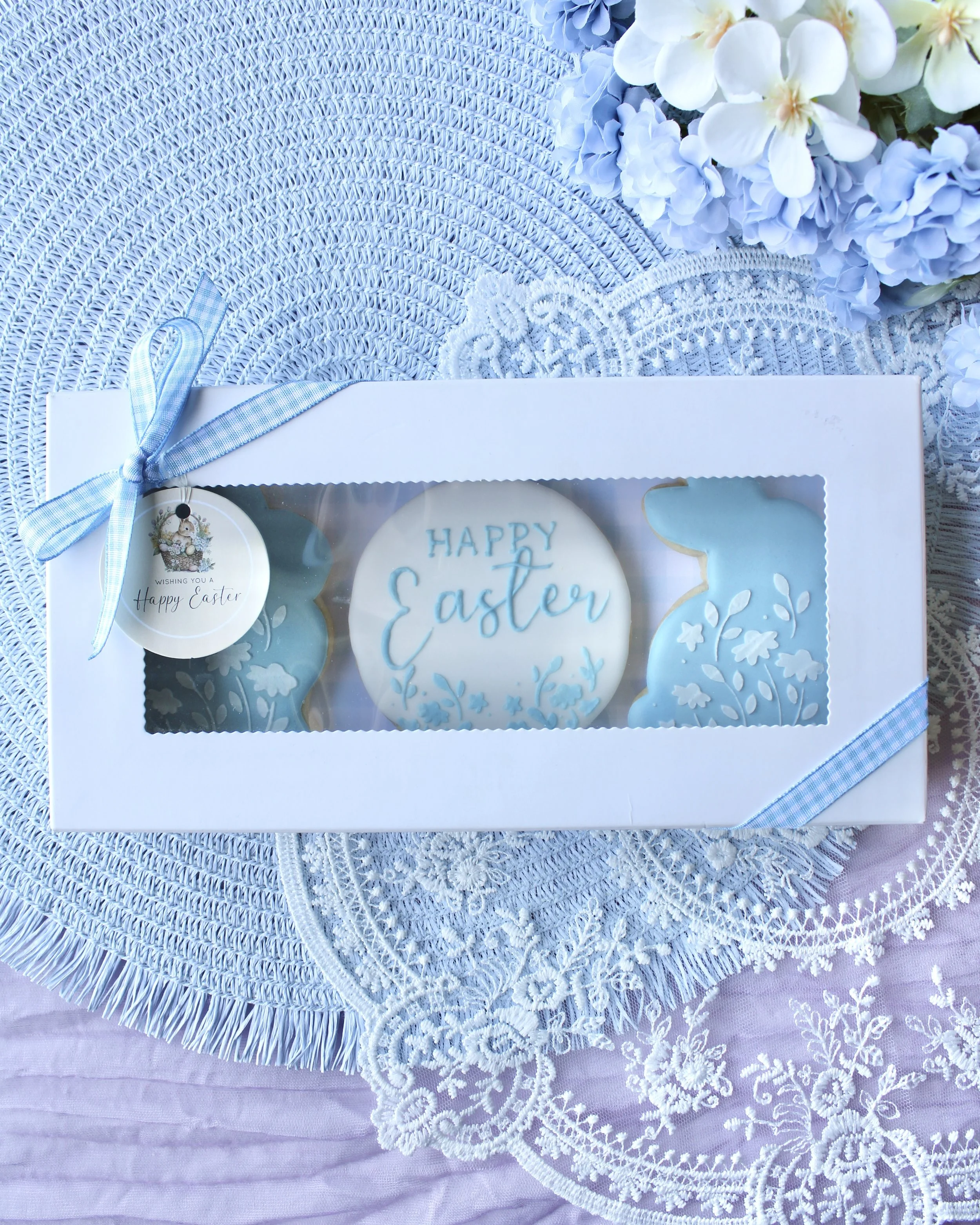 Easter-themed cookies in a white box with a blue ribbon, a decorative tag, and floral and lace decorations, set on a textured, circular placemat with a pastel purple tablecloth underneath.