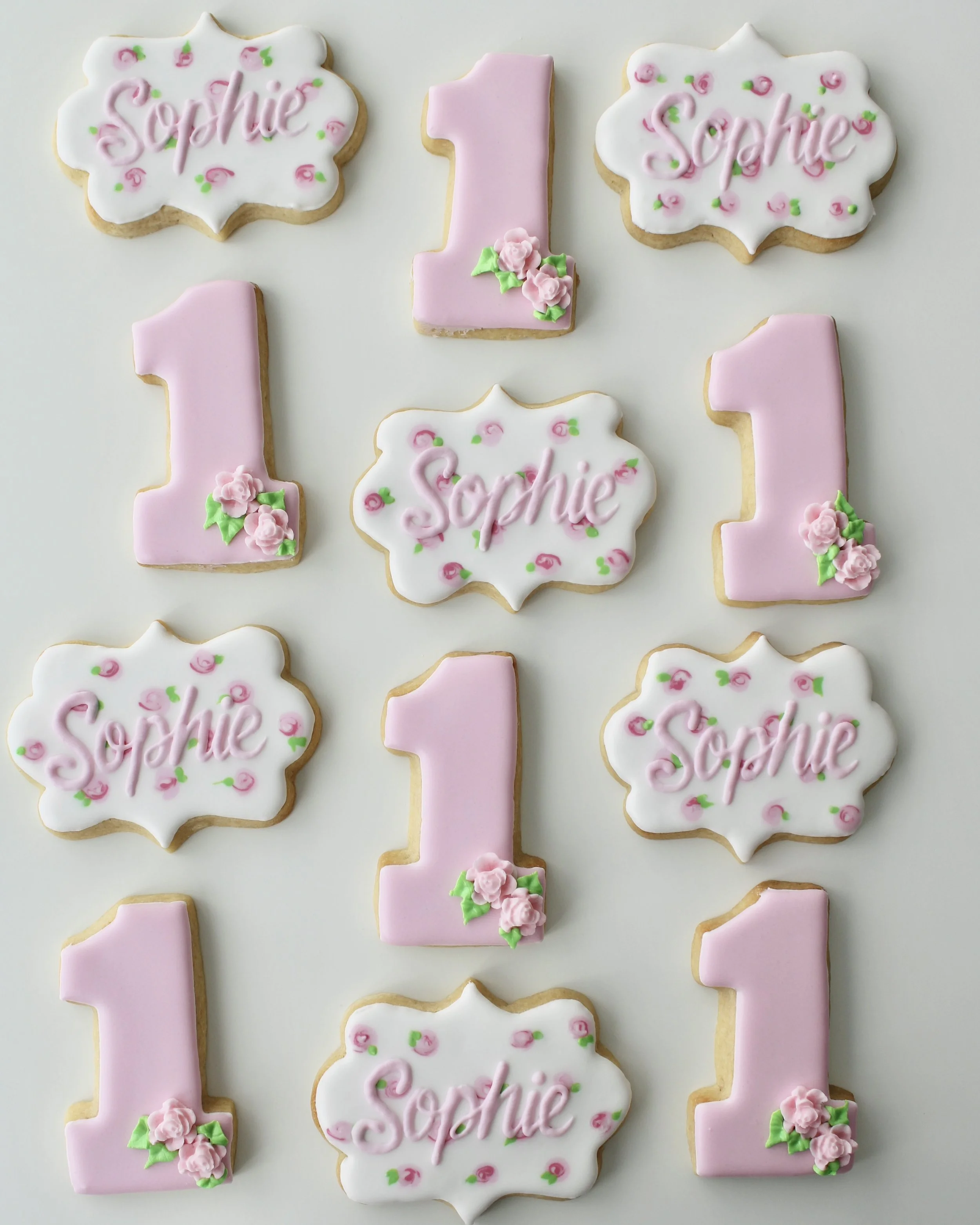 Custom first birthday cookies featuring pink number 1 cookies decorated with roses and white plaque cookies with the name “Sophie” in pink lettering and delicate floral details.