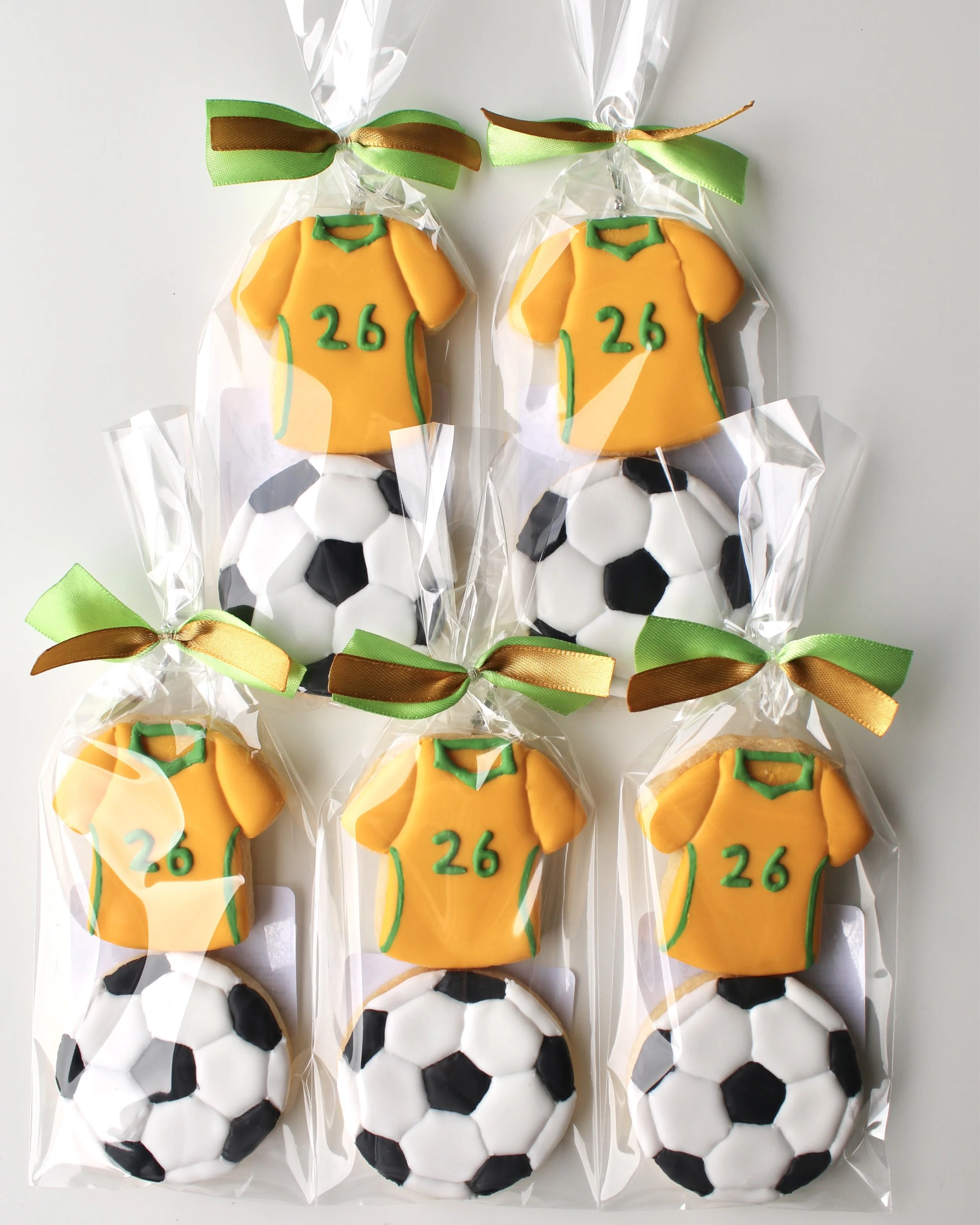 Close up of 5 cookie packs with two mini cookies each: Socceroos style jersey cookie with green accents and 26 detail, and soccer ball cookie in clear bags with ribbon.