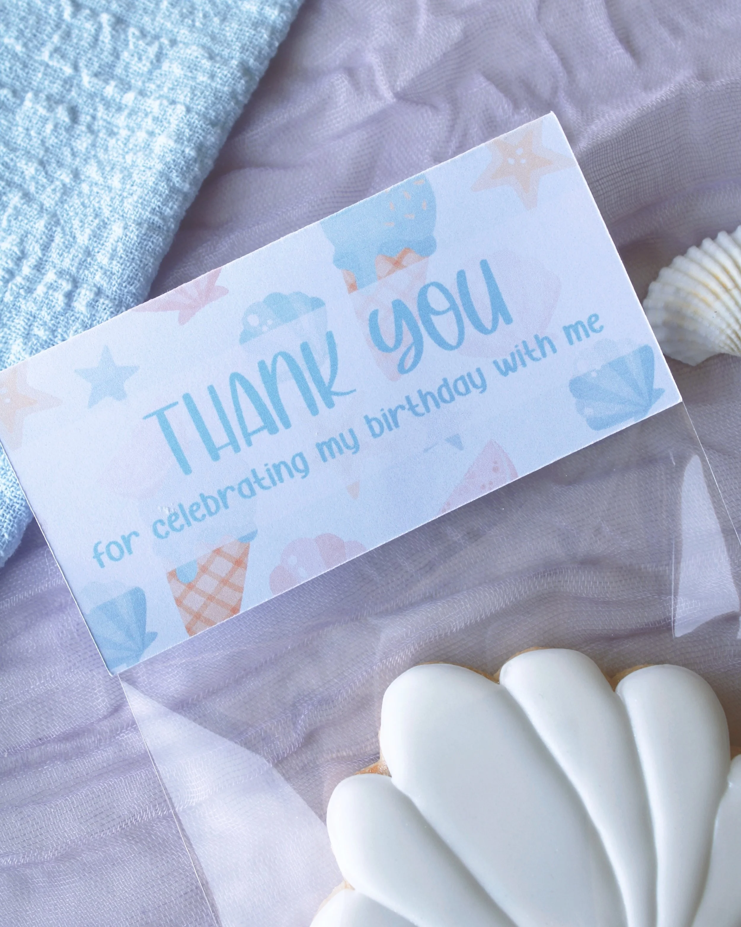 Close-up of seashell birthday party favour cookie packaging with a mermaid-themed “thank you for celebrating my birthday with me” topper on purple crinkled fabric.