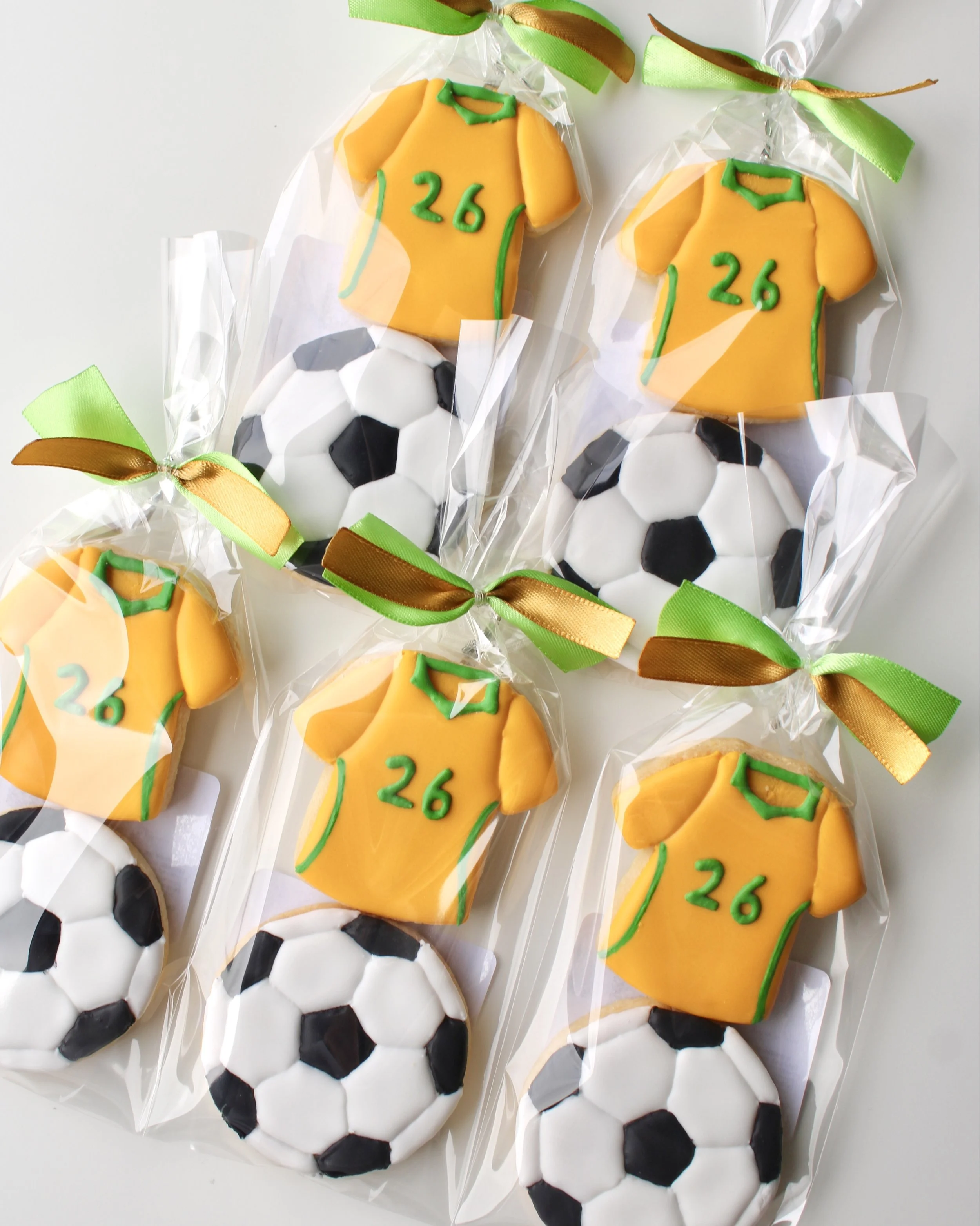 Close up of 5 cookie packs with two mini cookies each: Socceroos style jersey cookie with green accents and 26 detail, and soccer ball cookie in clear bags with ribbon.