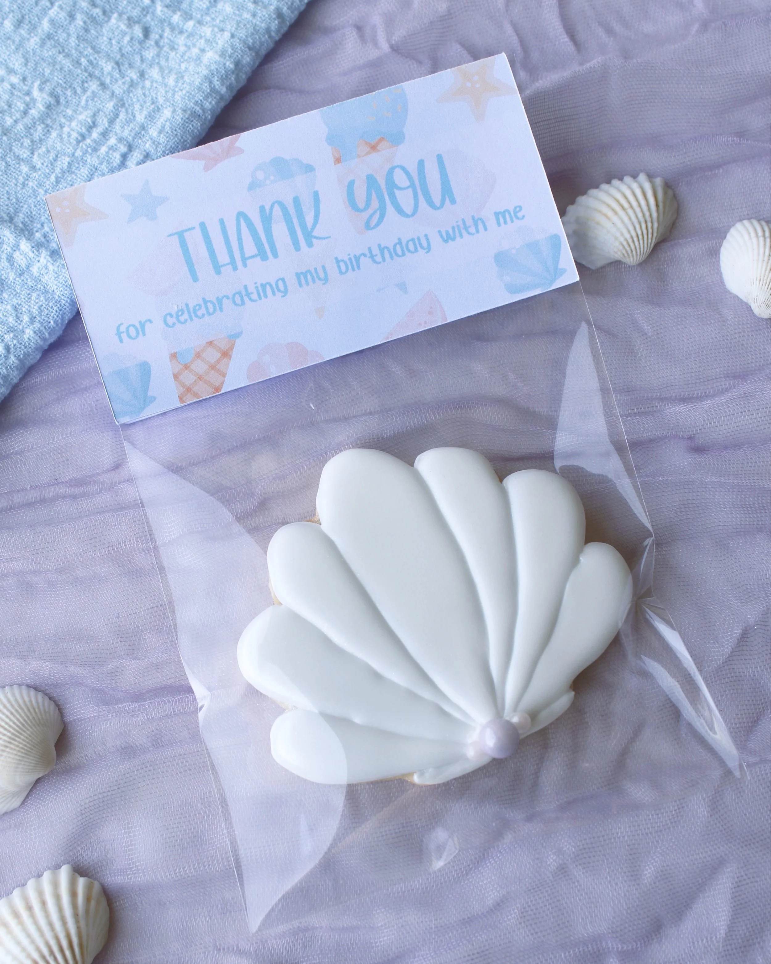 Seashell birthday party favour cookie in shimmering white with pearl details, packaged with a mermaid-themed “thank you for celebrating my birthday with me” topper on purple crinkled fabric.