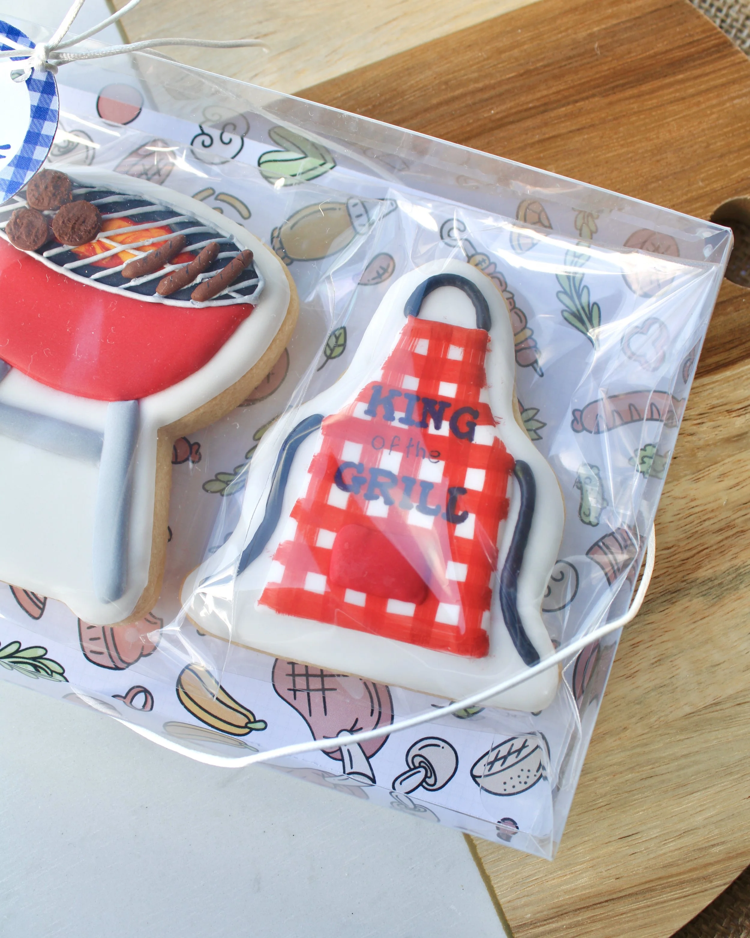Close up of Father's Day cookie featuring a red and white gingham patterned apron with "King of the Grill" written on it, styled on a wooden board.