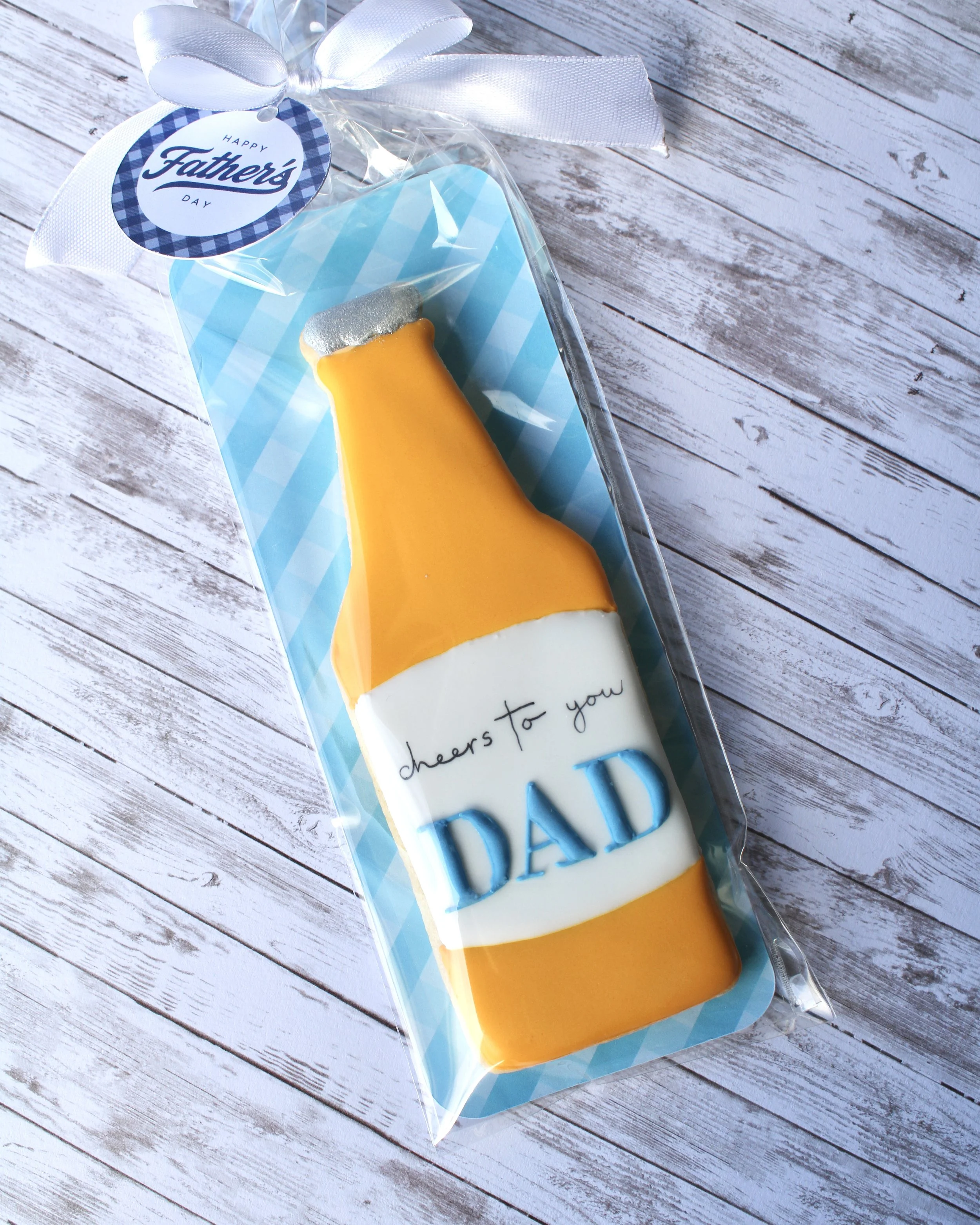 Father's Day beer bottle cookie decorated with golden yellow and white icing, featuring "Cheers to You Dad" label, blue gingham backer, white ribbon and gift tag on white washed grey wood background.