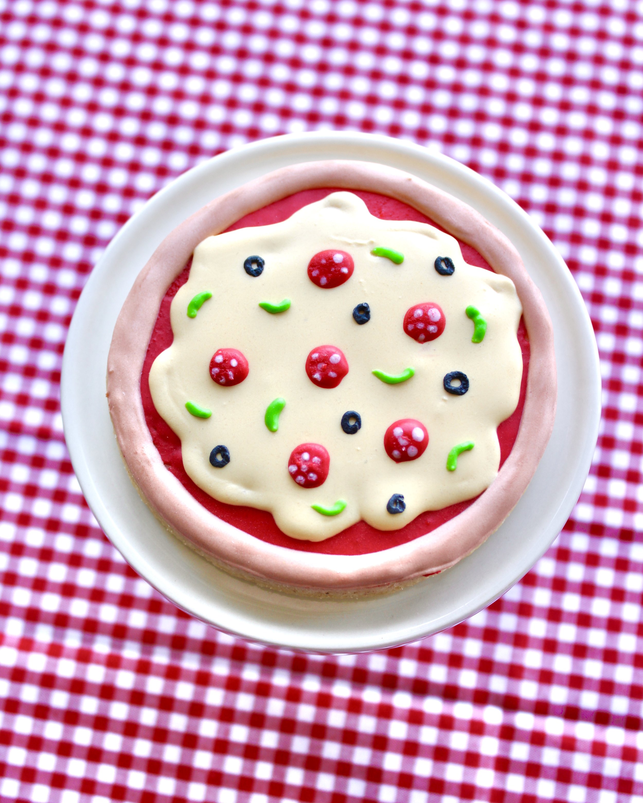Pizza-themed decorated cookie styled on a mini white cake stand with a red and white checkered fabric background.