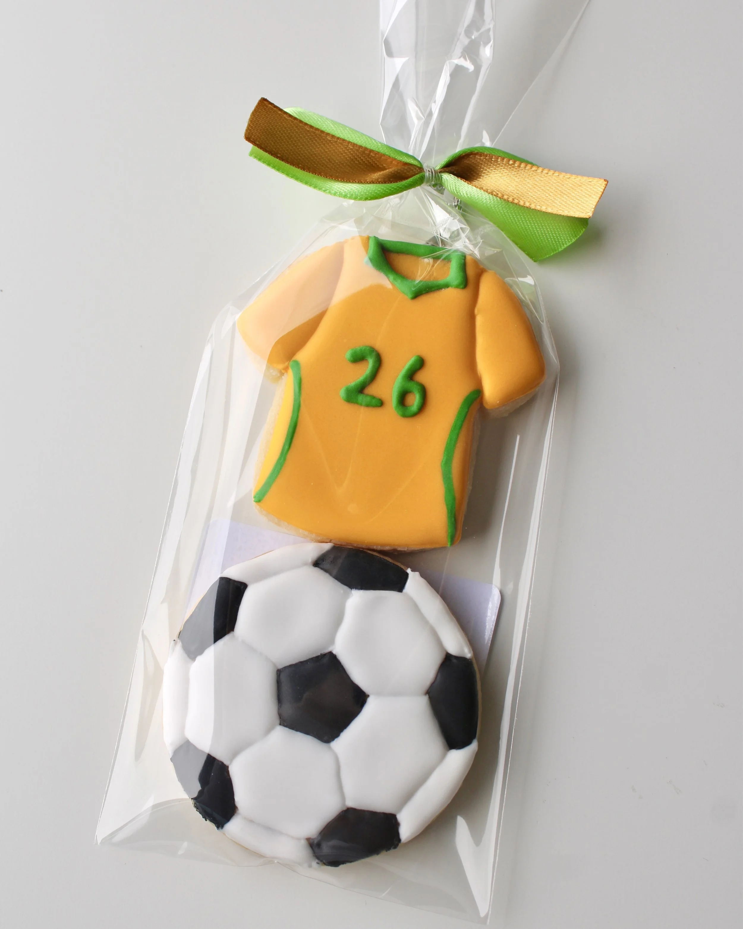Close up of cookie pack with two mini cookies: Socceroos style jersey cookie with green accents and 26 detail, and soccer ball cookie in clear bag with ribbon.