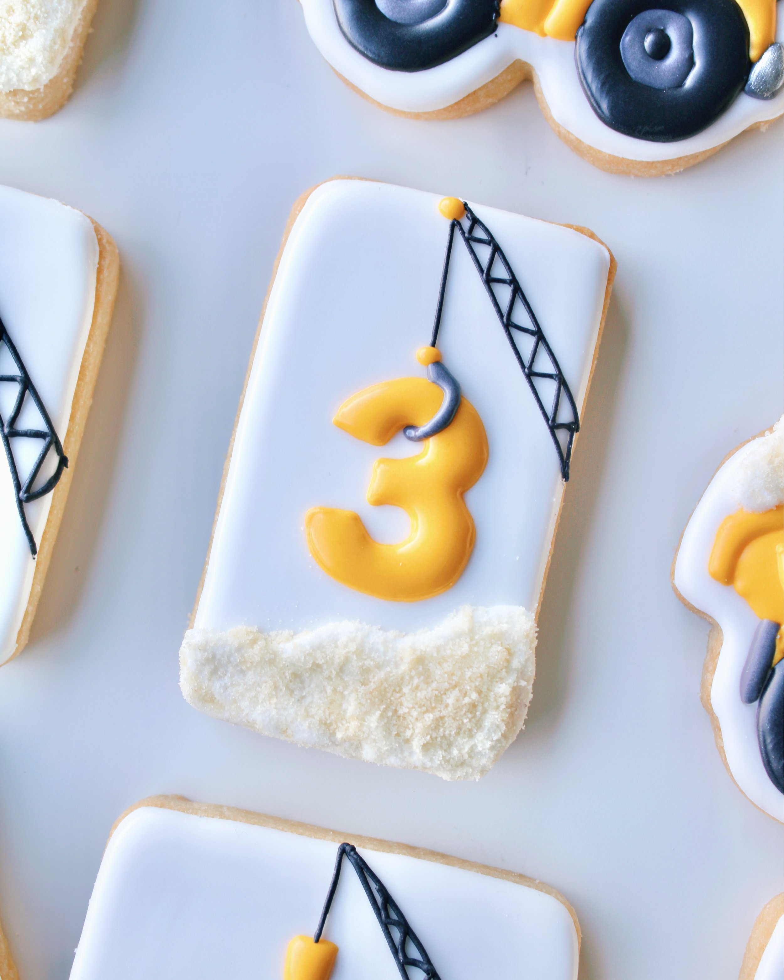 Close-up of a white rectangular cookie with a construction theme, featuring a crane holding a yellow number 3 with sand at the bottom.
