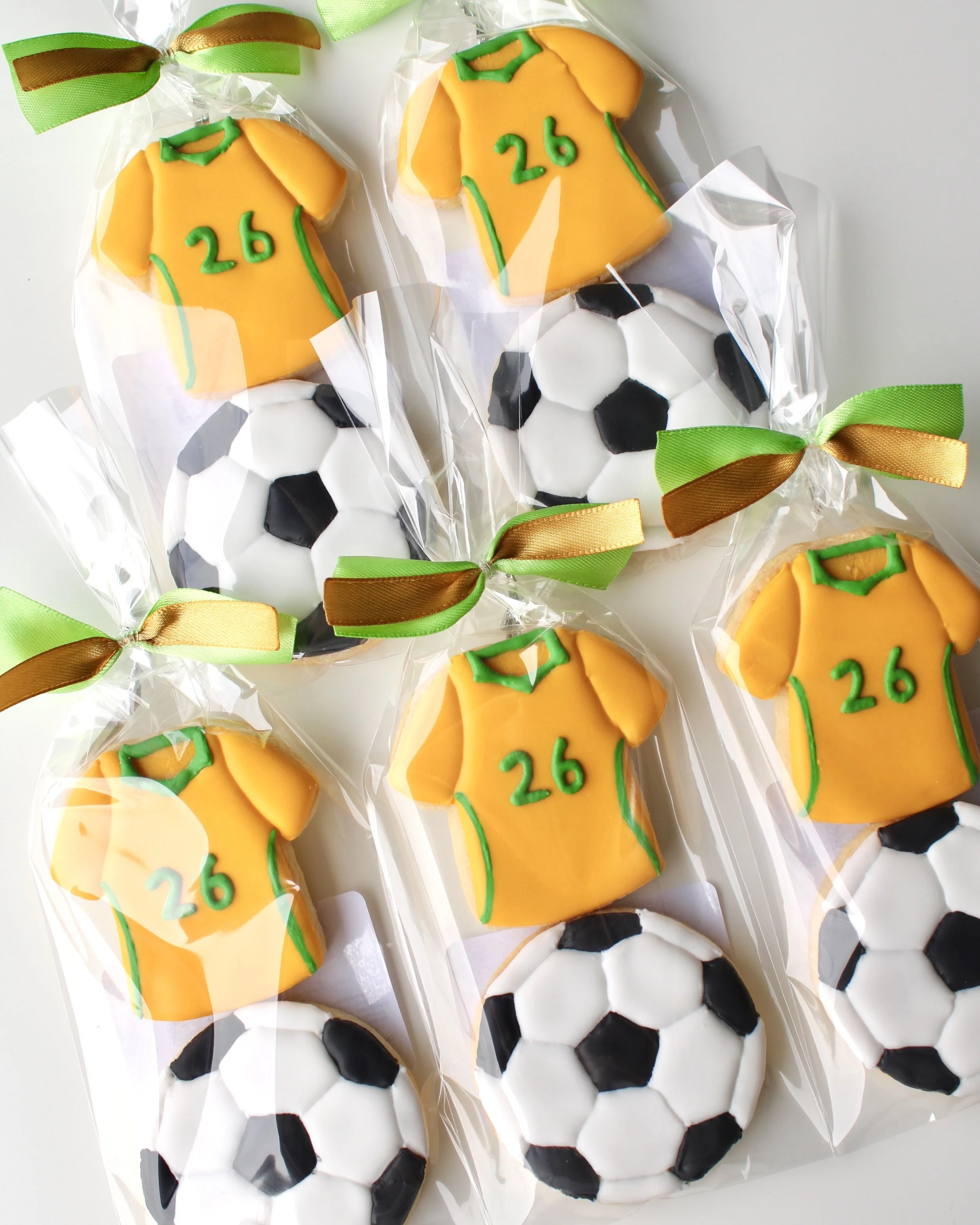 Close up of 5 cookie packs with two mini cookies each: Socceroos style jersey cookie with green accents and 26 detail, and soccer ball cookie in clear bags with ribbon.