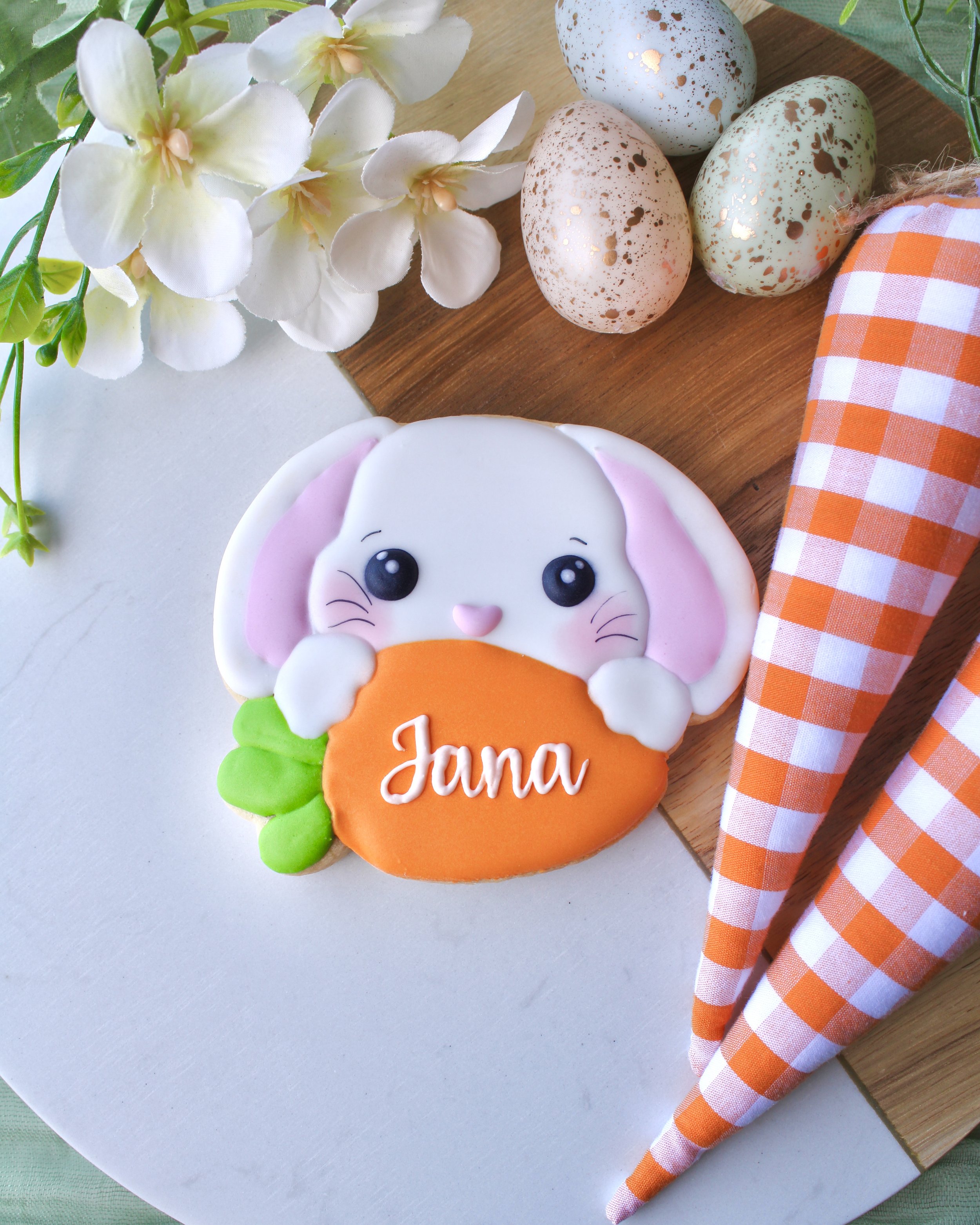 Personalised Easter bunny sugar cookie with white face, pink ears and carrot, “Jana” in icing script, styled on wood and marble board with gingham carrots, eggs and spring flowers.
