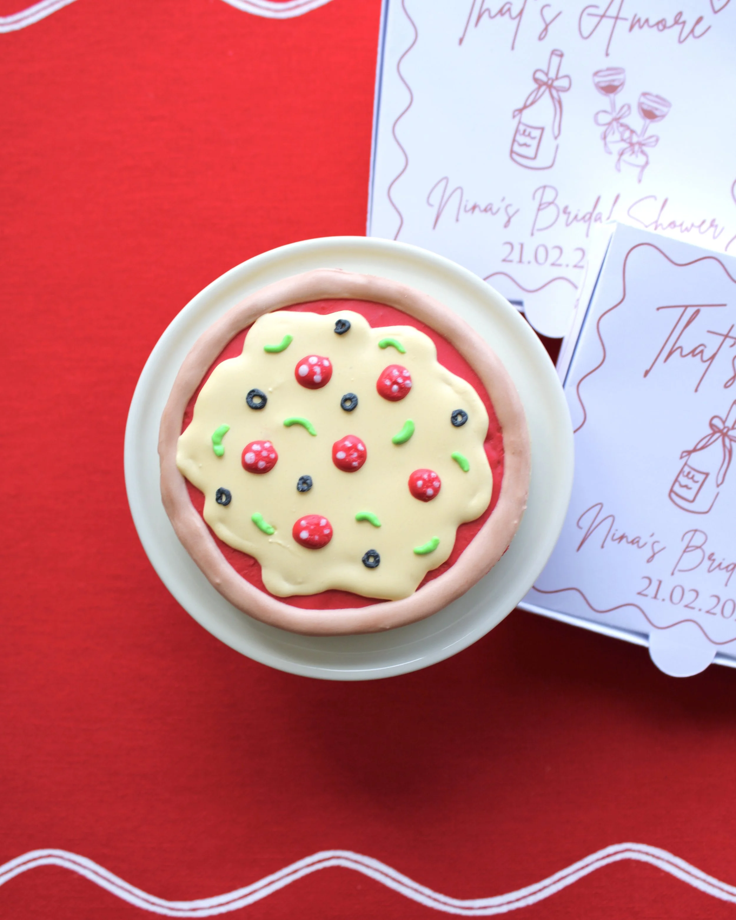 Close-up of a mini pizza cookie box on a mini cake stand, with two additional mini pizza cookie boxes in the background, styled on red fabric.