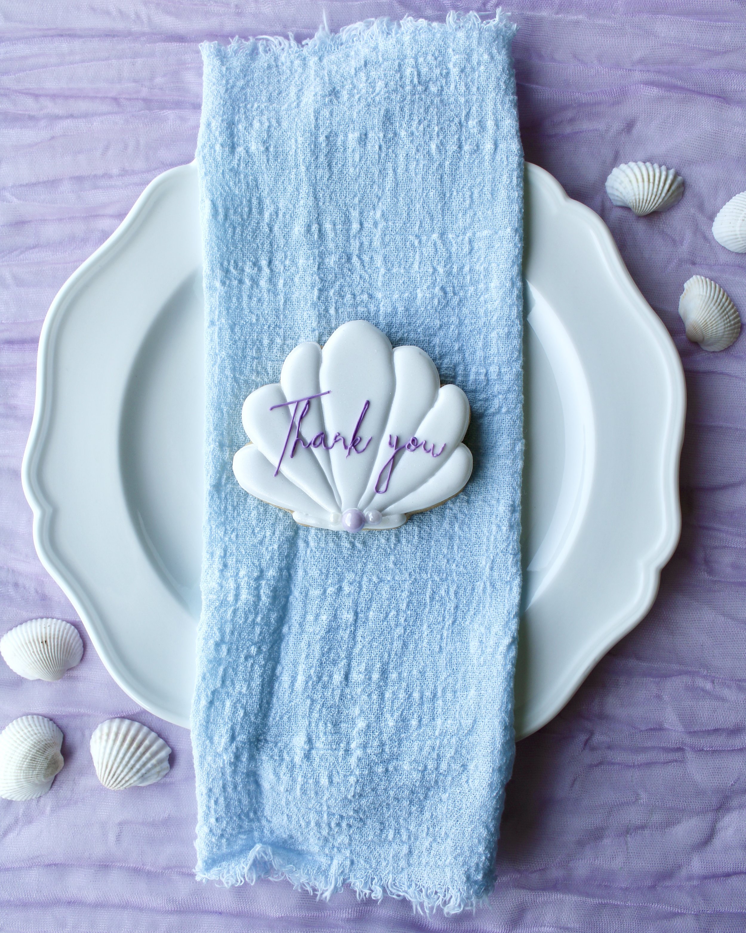 White seashell cookie with “thank you” written in purple cursive icing, placed on a white vintage plate over blue linen napkin and purple crinkled fabric with scattered seashells.