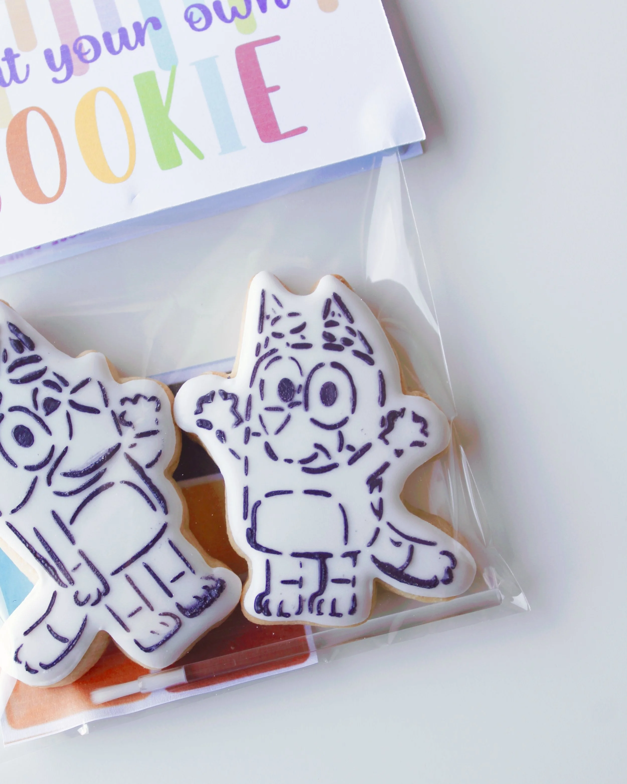 Paint your own cookie pack featuring Bluey and Bingo cookies, white icing designs with black stencilled outlines, and a rainbow “Paint Your Own” bag topper.