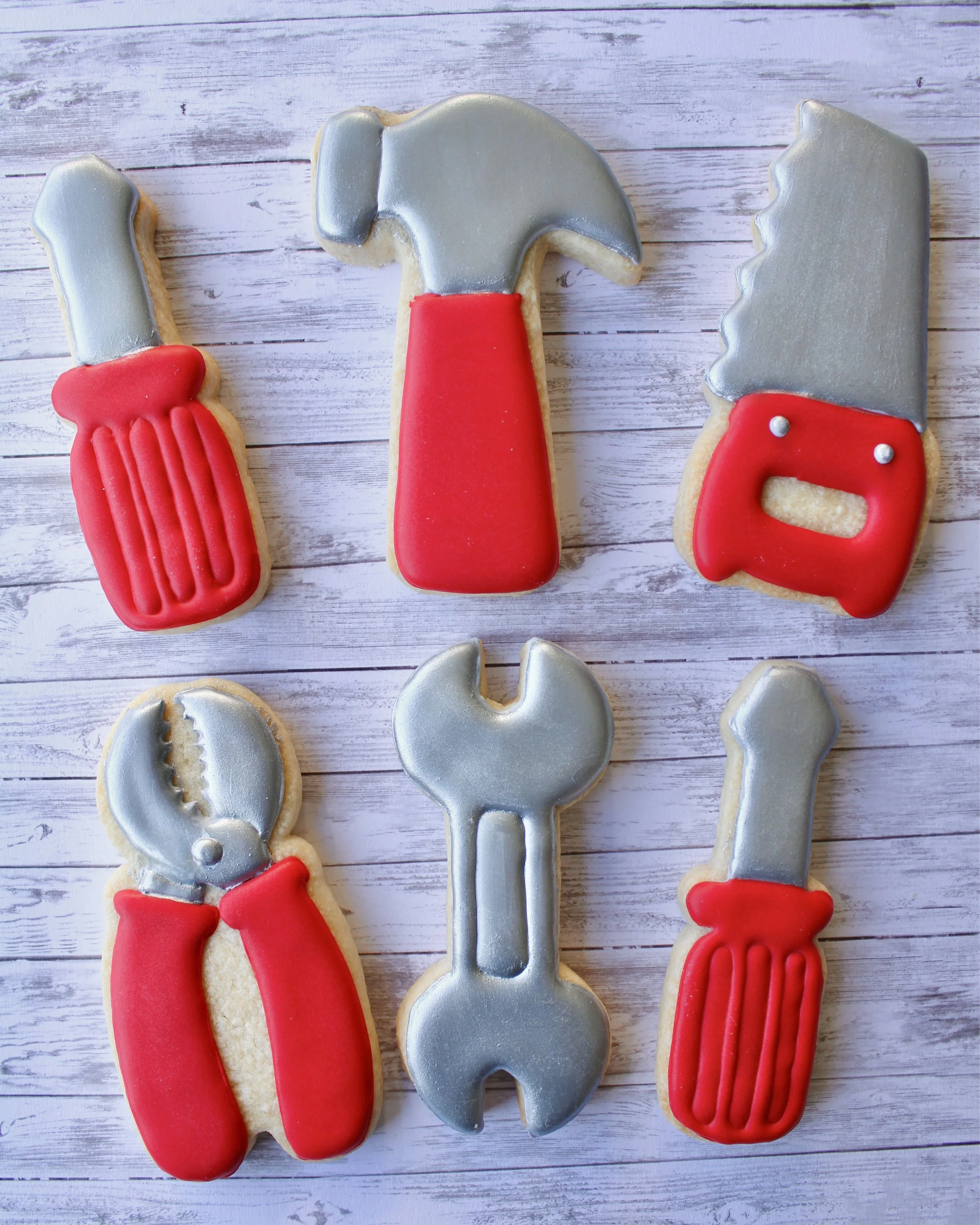 Father's Day cookie tools decorated in silver and red icing, featuring two screwdrivers, a hammer, saw, pliers and a wrench, styled on a grey washed wooden background.