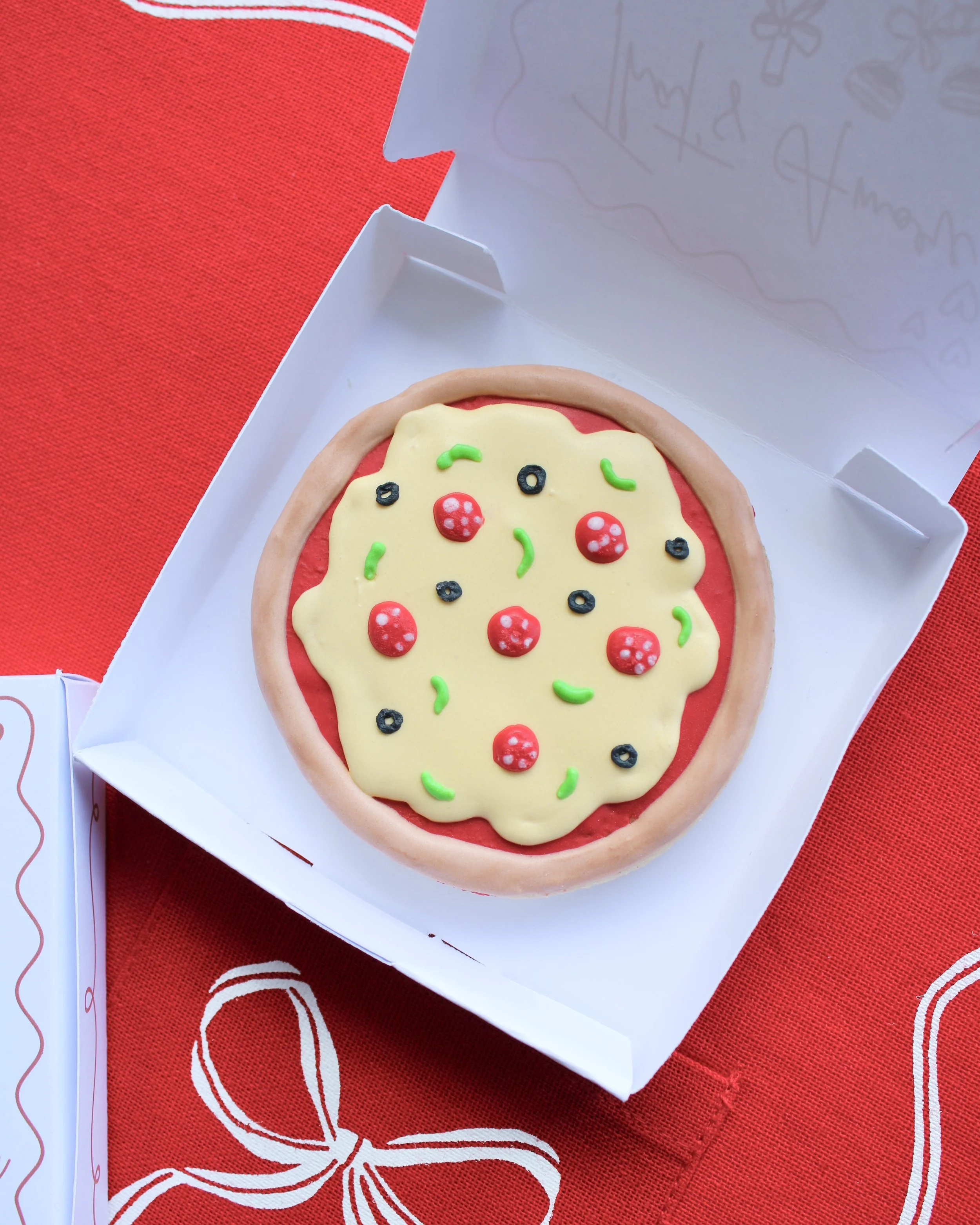 Close-up of a mini pizza cookie box opened to reveal a mini pizza cookie, styled on red fabric.