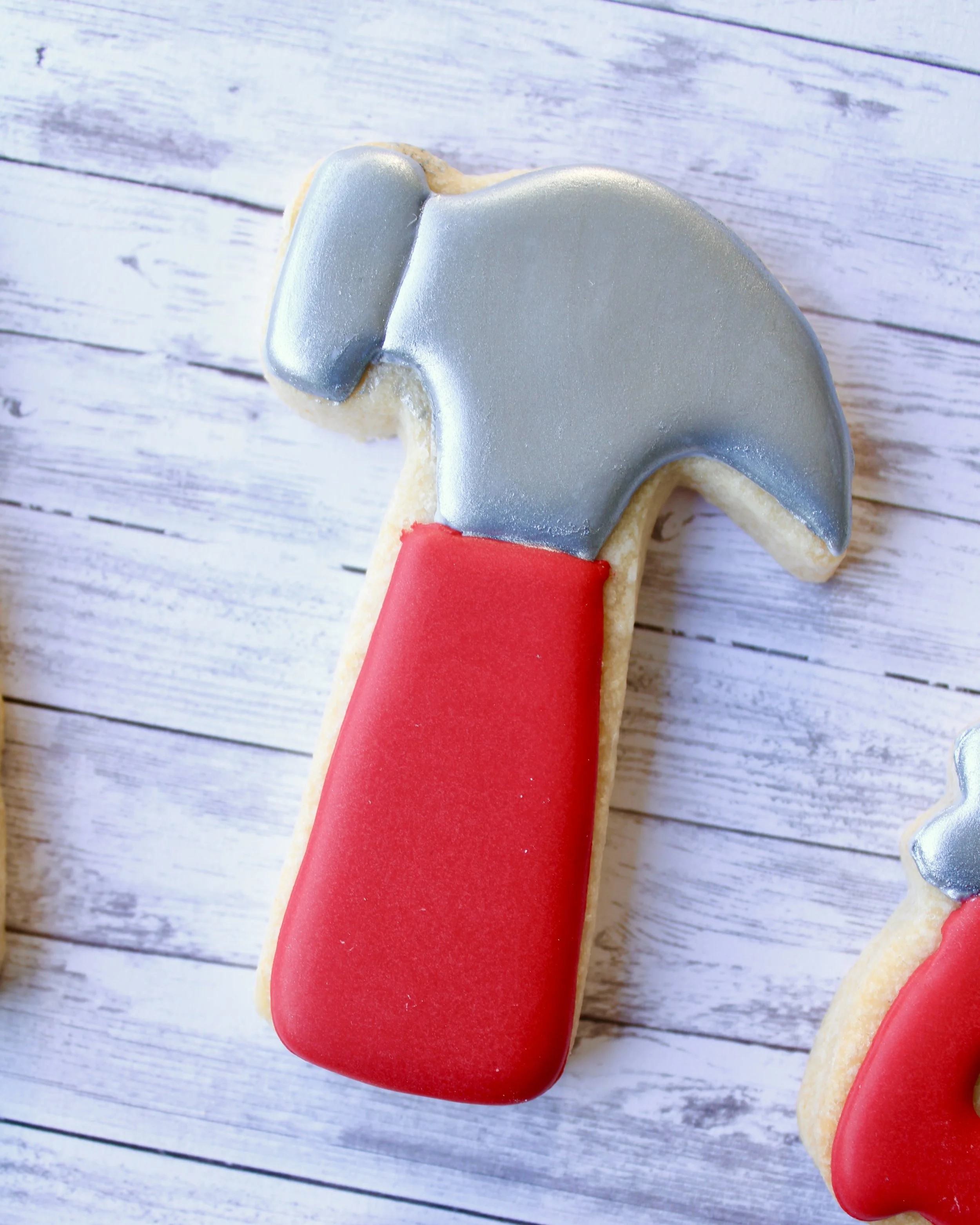 Close up of Father's Day tool cookie shaped like a hammer, with silver icing and a red icing handle, styled on a grey washed wooden background.
