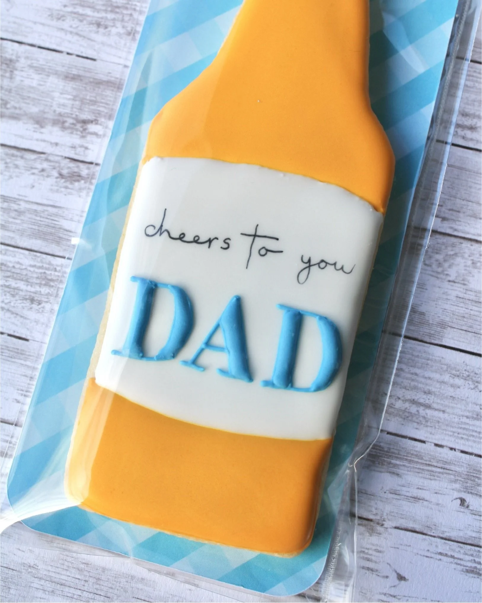 Close up of Father's Day beer bottle cookie decorated with golden yellow and white icing, featuring "Cheers to You Dad" label, with blue gingham backer and white washed grey wood background.