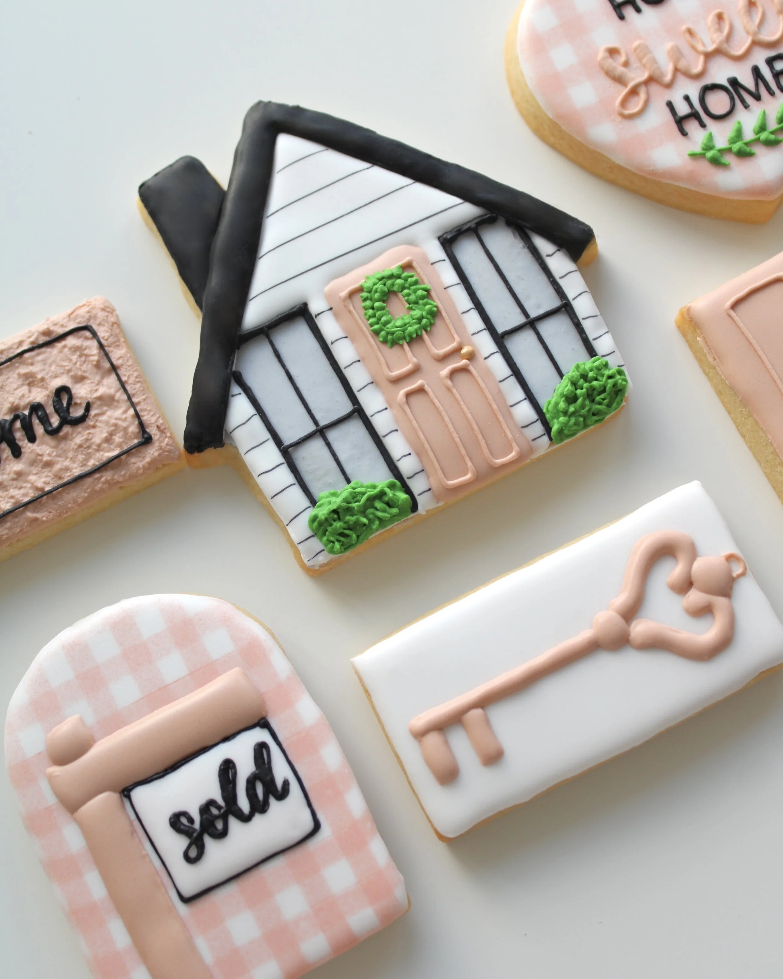 Flat lay of six New Home cookies in white, beige, black and green with beige gingham print, including a house, door, “Home Sweet Home” heart, sold sign, welcome mat and key.
