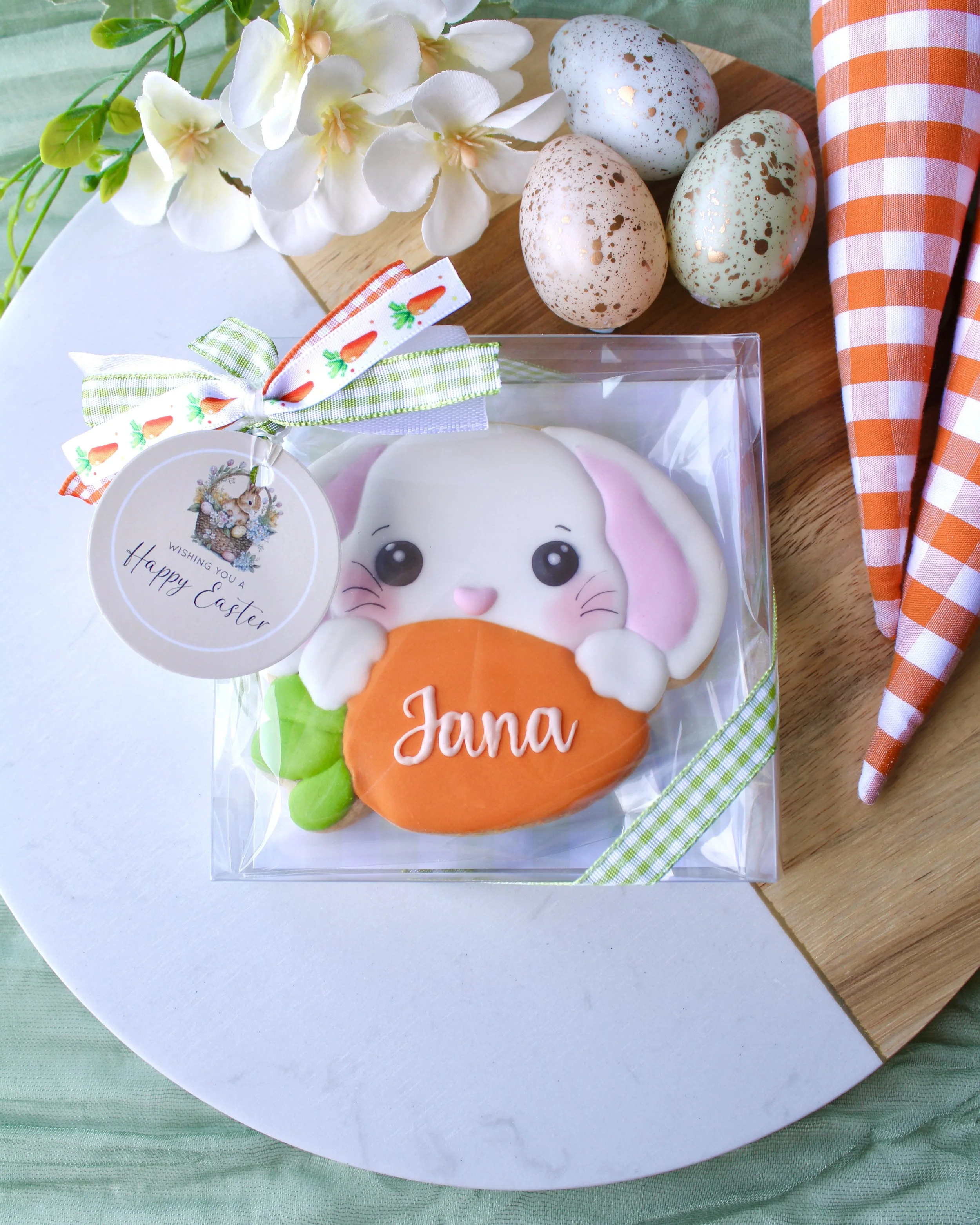 Personalised Easter bunny sugar cookie with “Jana”, in a clear gift box with Happy Easter tag and gingham ribbon, styled on wood and marble board with eggs and spring flowers.