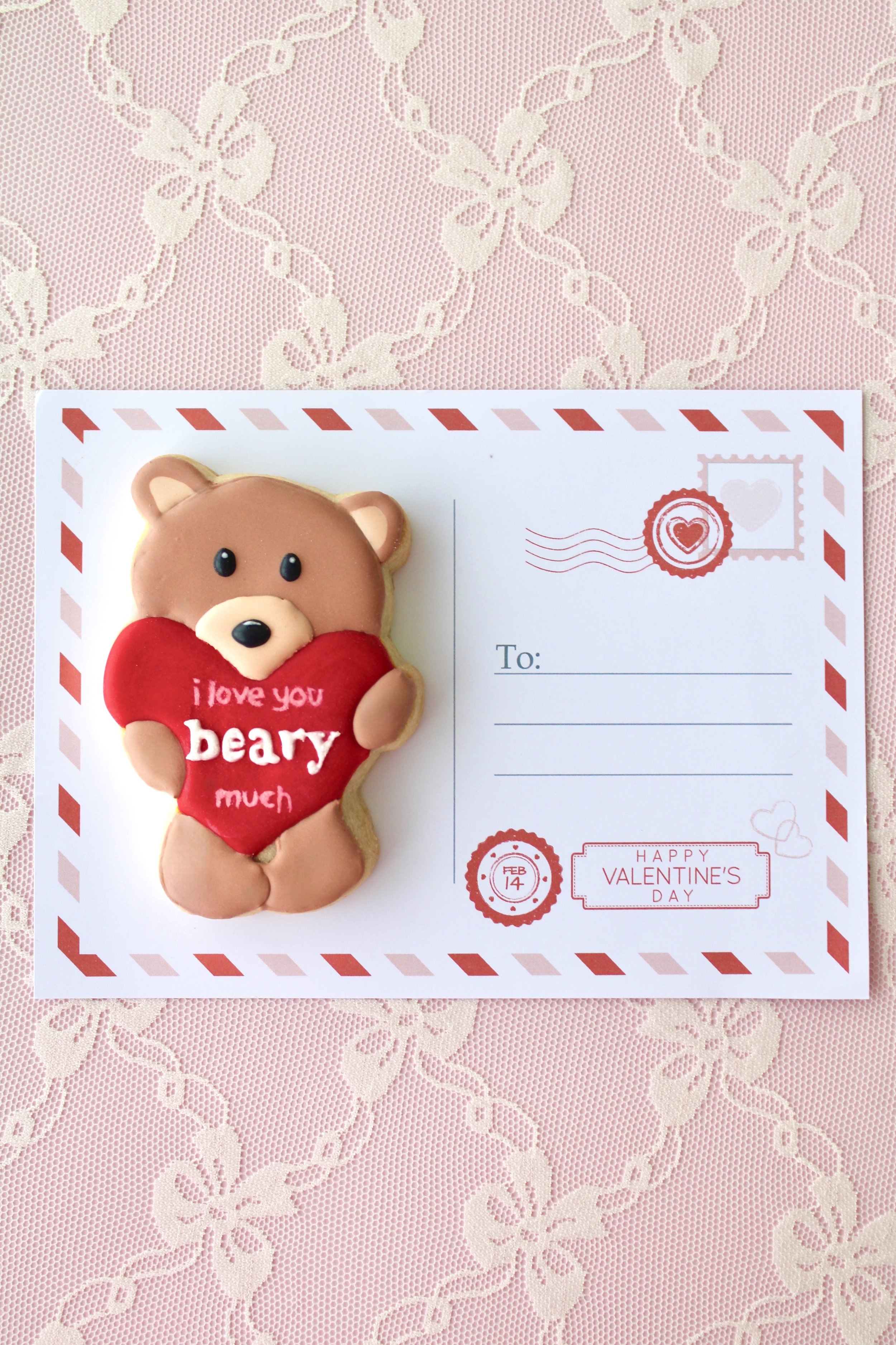 Brown bear cookie holding a red heart with the words “I love you beary much” in white script, styled on a vintage Valentine postcard with red and pink envelope border and stamps, on pink and white lace fabric with lace bows.