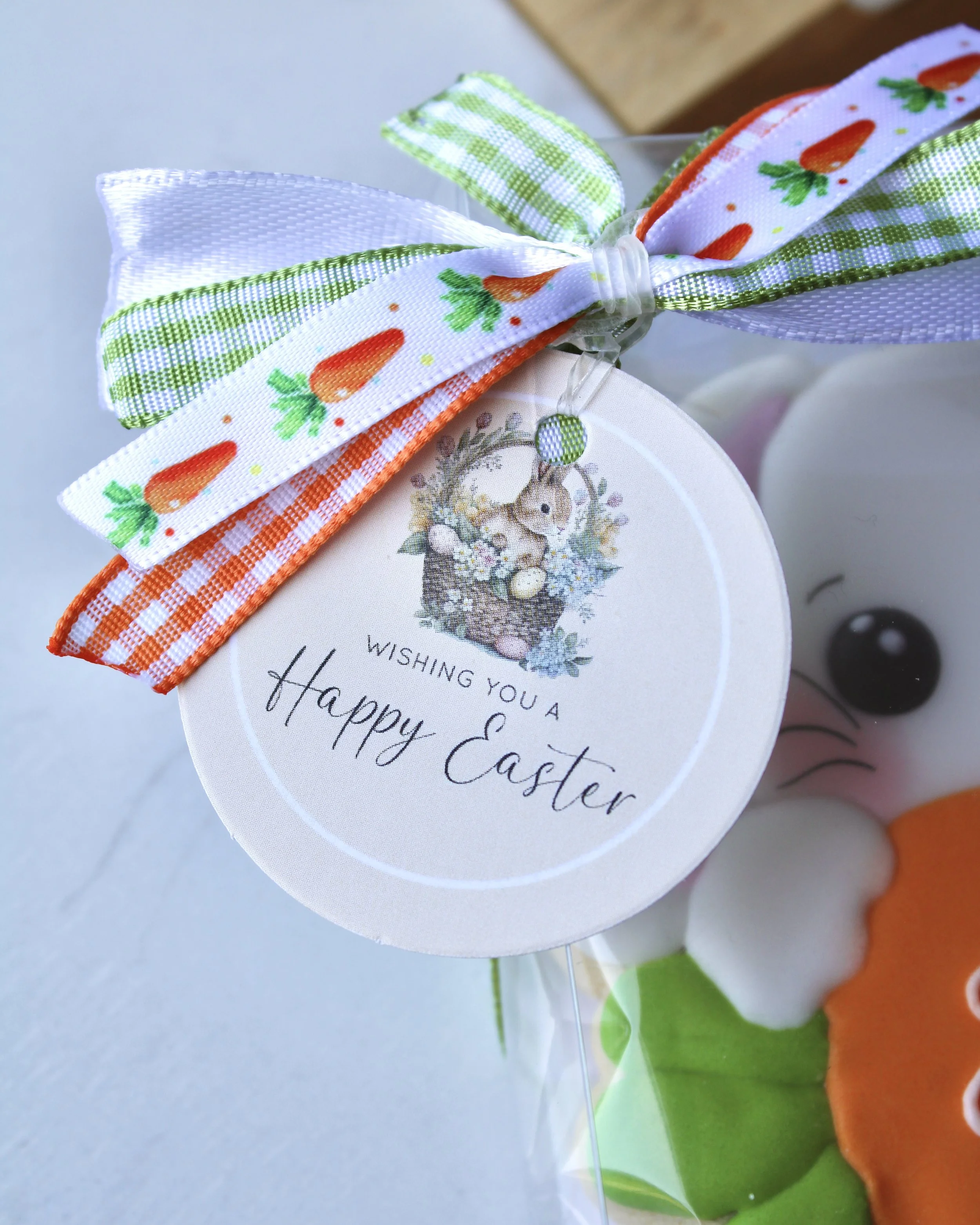 Close-up Easter gift tag with bunny in a basket and “Wishing You a Happy Easter,” tied to a cookie box with a four-ribbon bow in carrot, white, green and orange patterns.