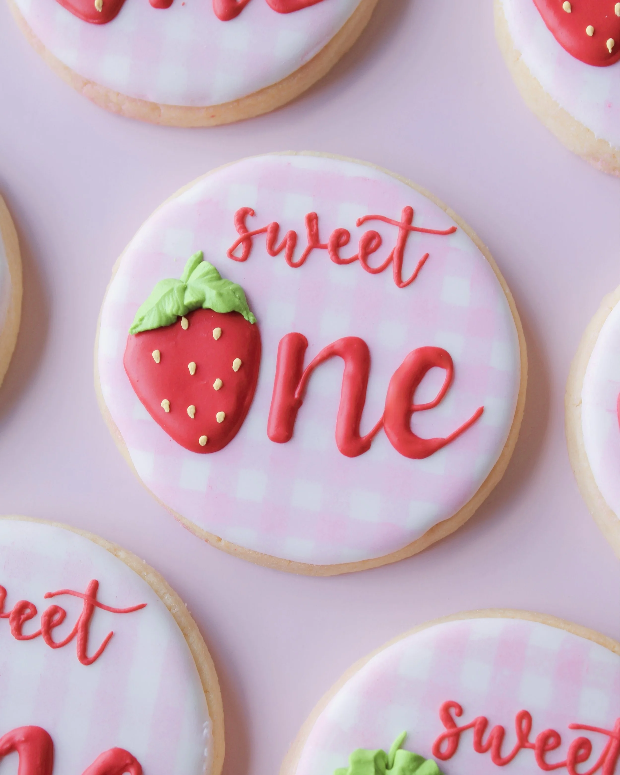Close-up of “Sweet One” themed cookie with pink and white gingham icing and “one” written in red icing, featuring a strawberry-shaped “o”.