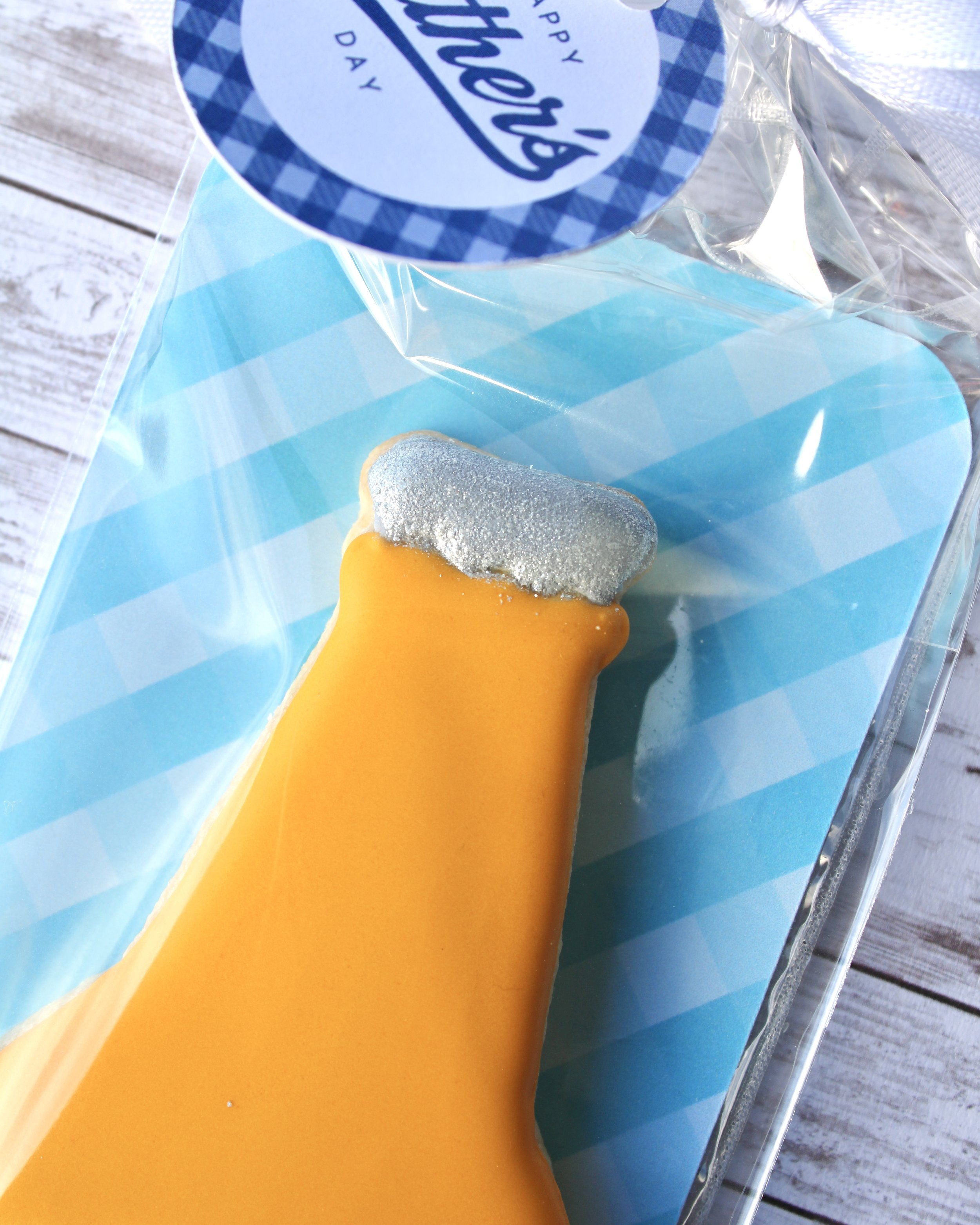Close up of Father's Day beer bottle cookie with silver edible paint bottle cap and golden yellow icing, in a clear cookie bag with blue gingham backer, styled on a white washed grey wood background.