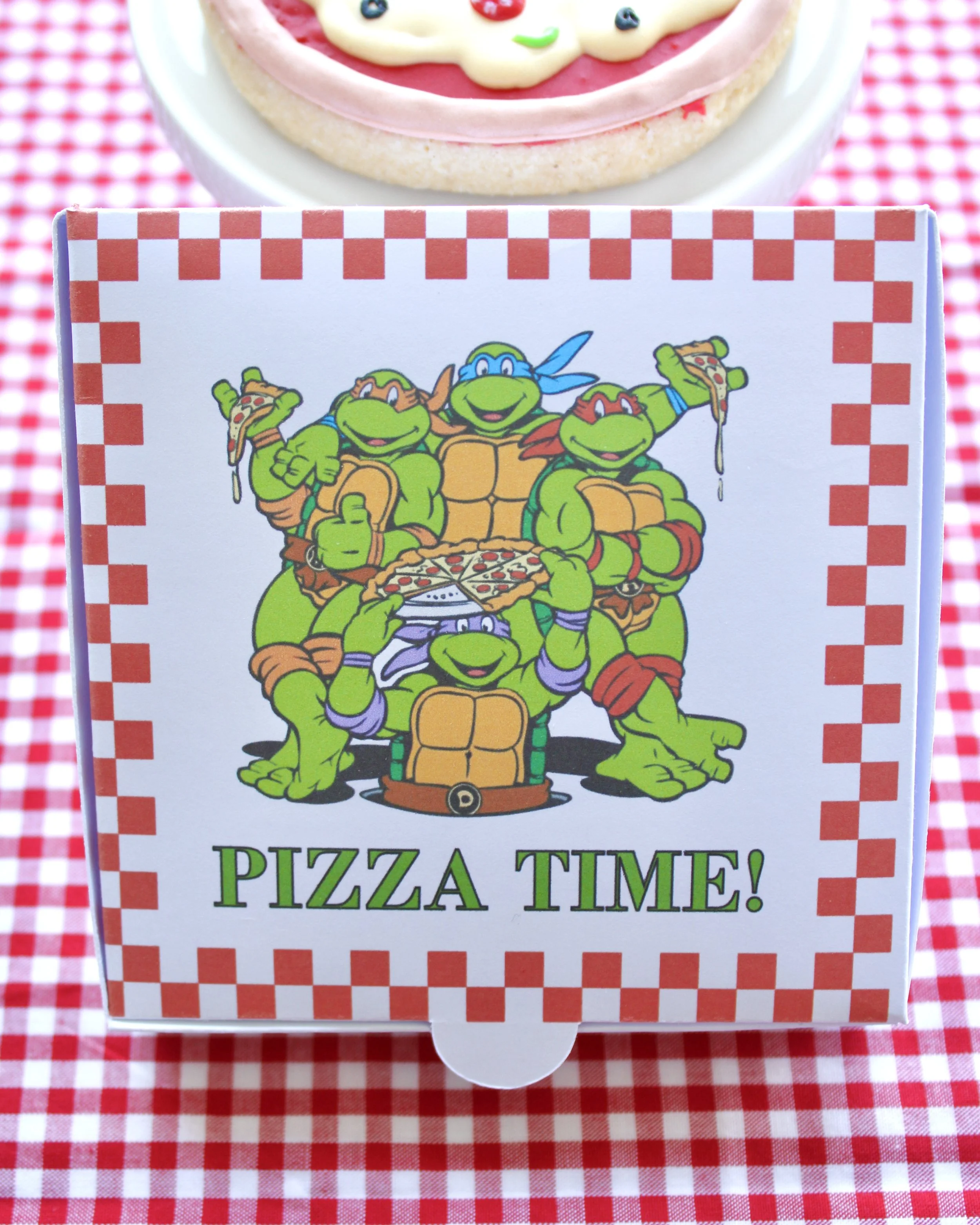 Mini pizza box featuring Teenage Mutant Ninja Turtles cookies on a red and white checkered background, with a pizza-shaped cookie in the background.