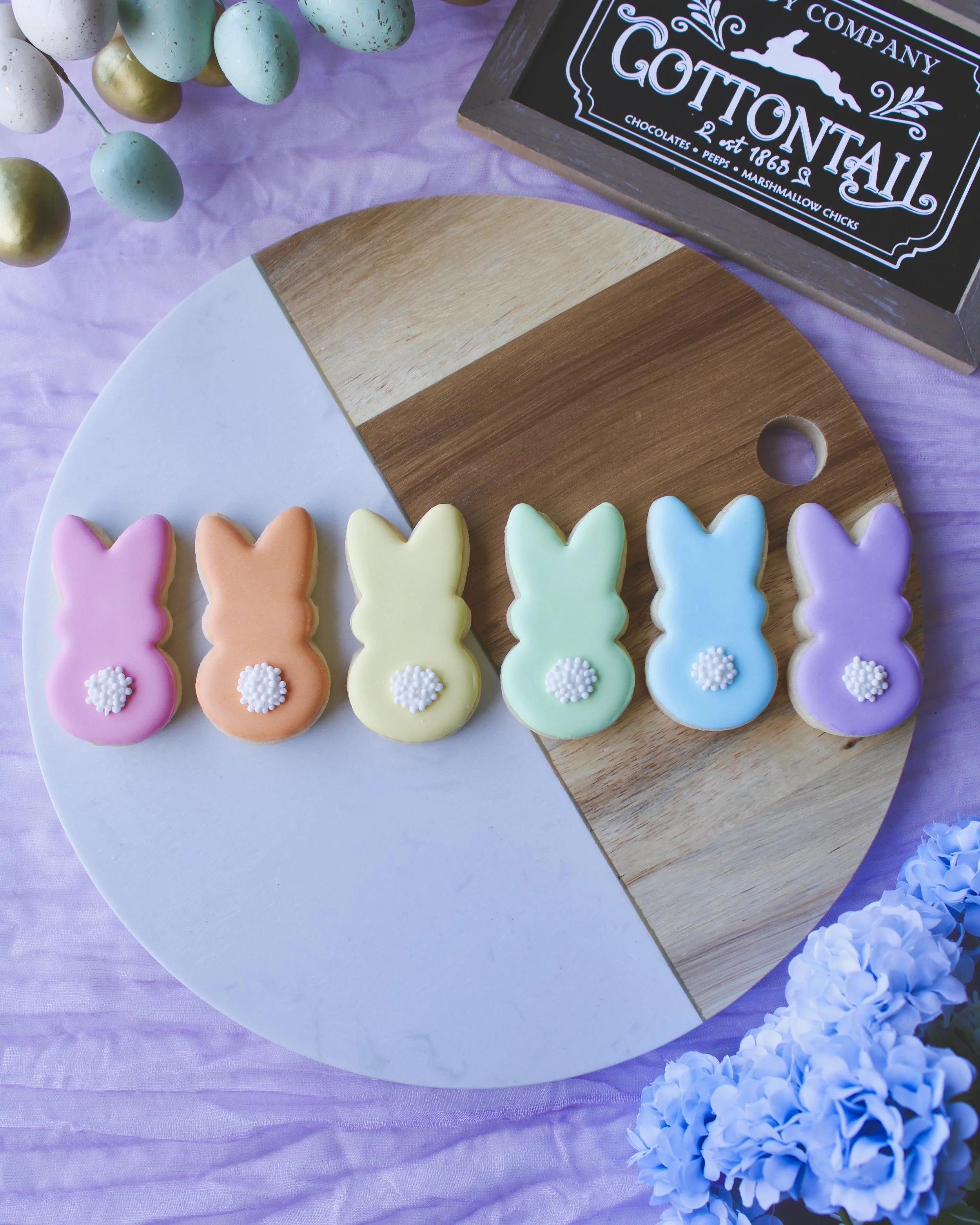 Six mini Easter Bunny Peeps cookies in rainbow pastel colours with white tails on a marble and wood board, styled with lilac fabric, Easter eggs, hydrangeas and a chalkboard sign.