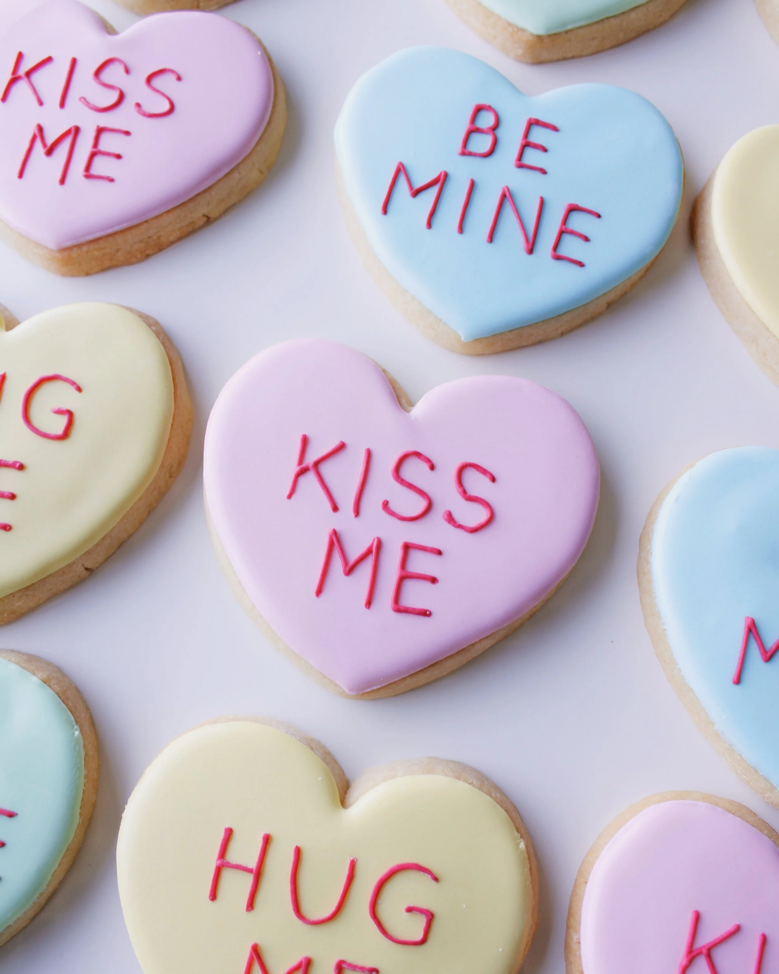 Close-up of a heart-shaped cookie decorated like a conversation heart candy in pastel pink icing with red text reading “kiss me.”