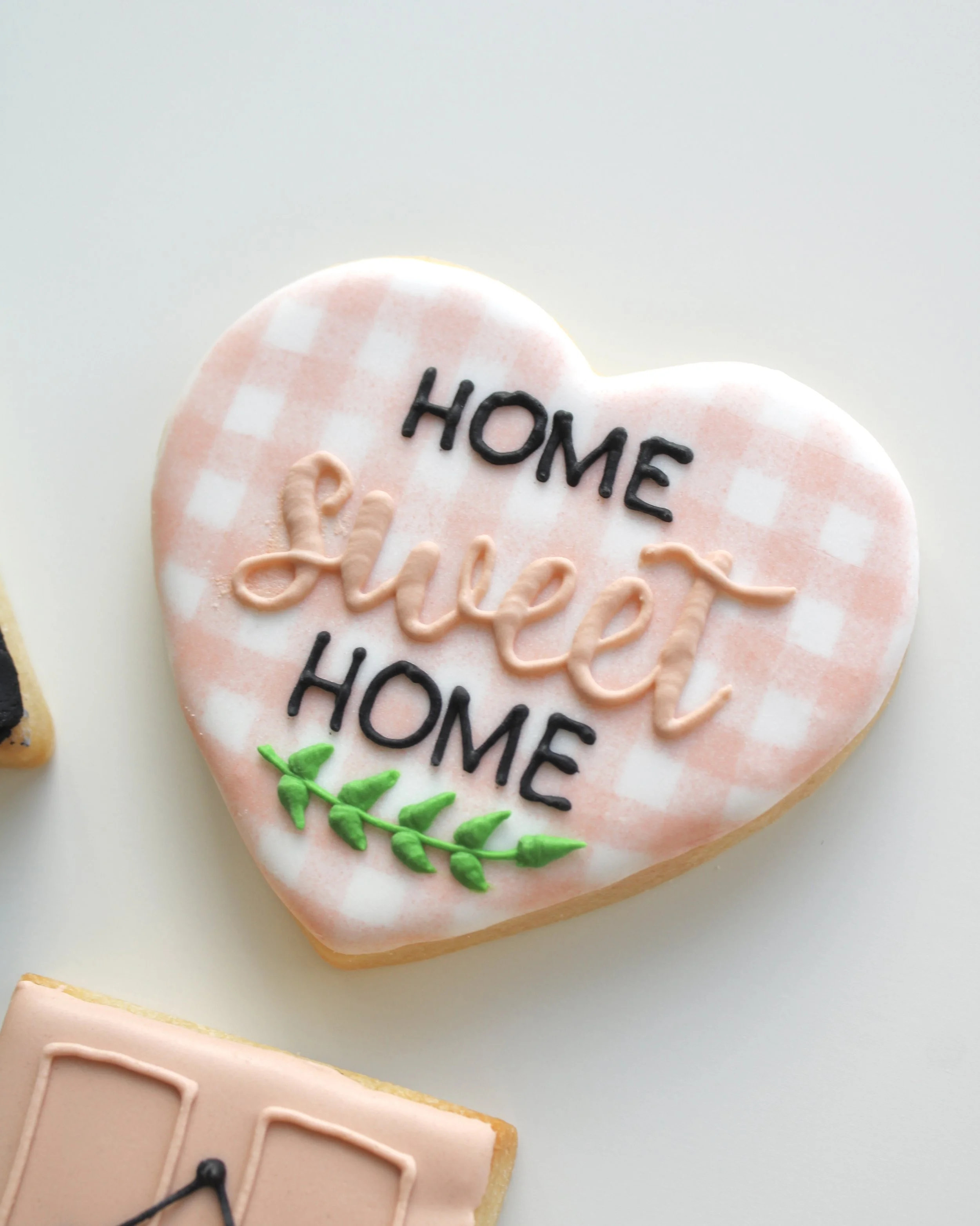 Close-up of a heart-shaped cookie iced in white with beige gingham detailing, featuring “home sweet home” written in black icing and a green leafy branch at the bottom.