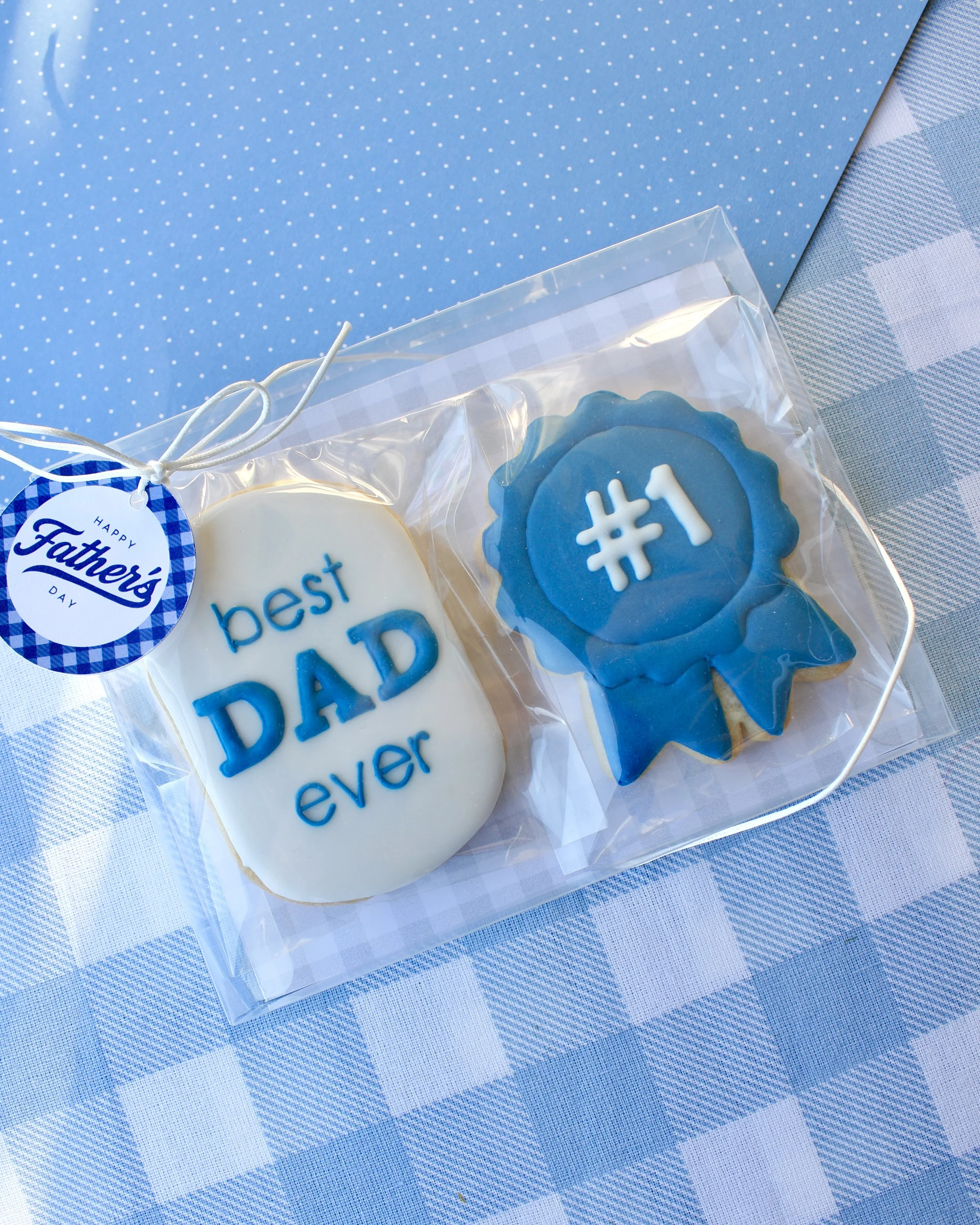 Father's Day cookie gift set featuring two handmade sugar cookies: a "Best Dad Ever" plaque with blue icing and a blue award ribbon cookie with "#1" in white icing, styled on blue gingham fabric.