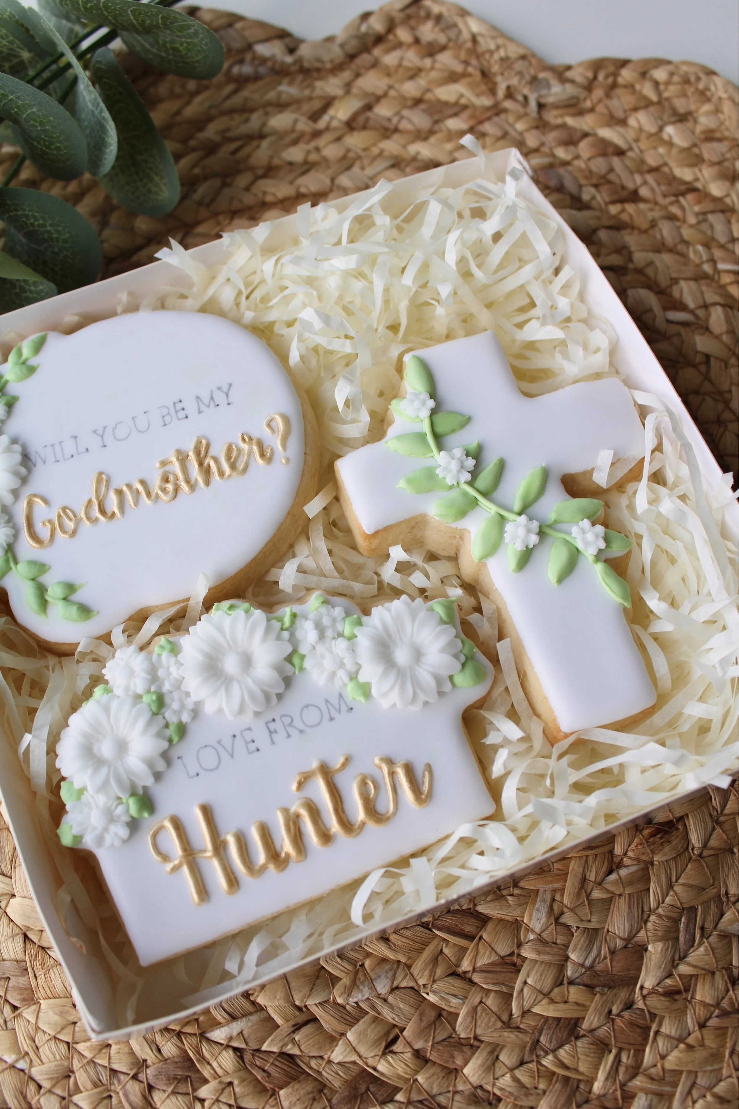 Godparent proposal cookie box with 3 white iced cookies: cross with green vine, “Will you be my Godmother?” cookie, and “Love From Hunter” cookie with gold text, green leaves, and white flowers.