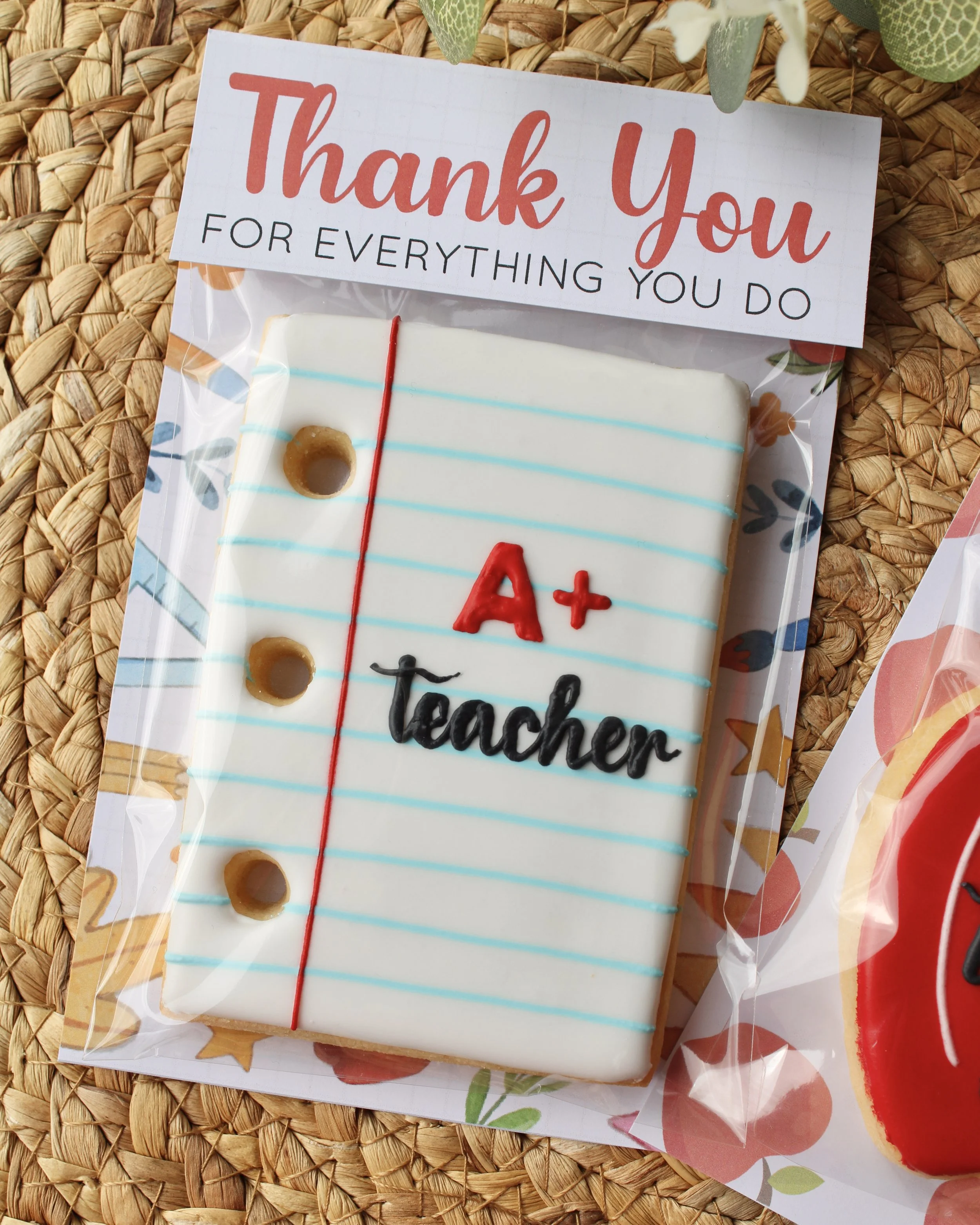 Individually packaged teacher cookie shaped like a notepad with “A+ teacher” icing and a “Thank you for everything you do” topper, styled with eucalyptus on a hyacinth placemat.