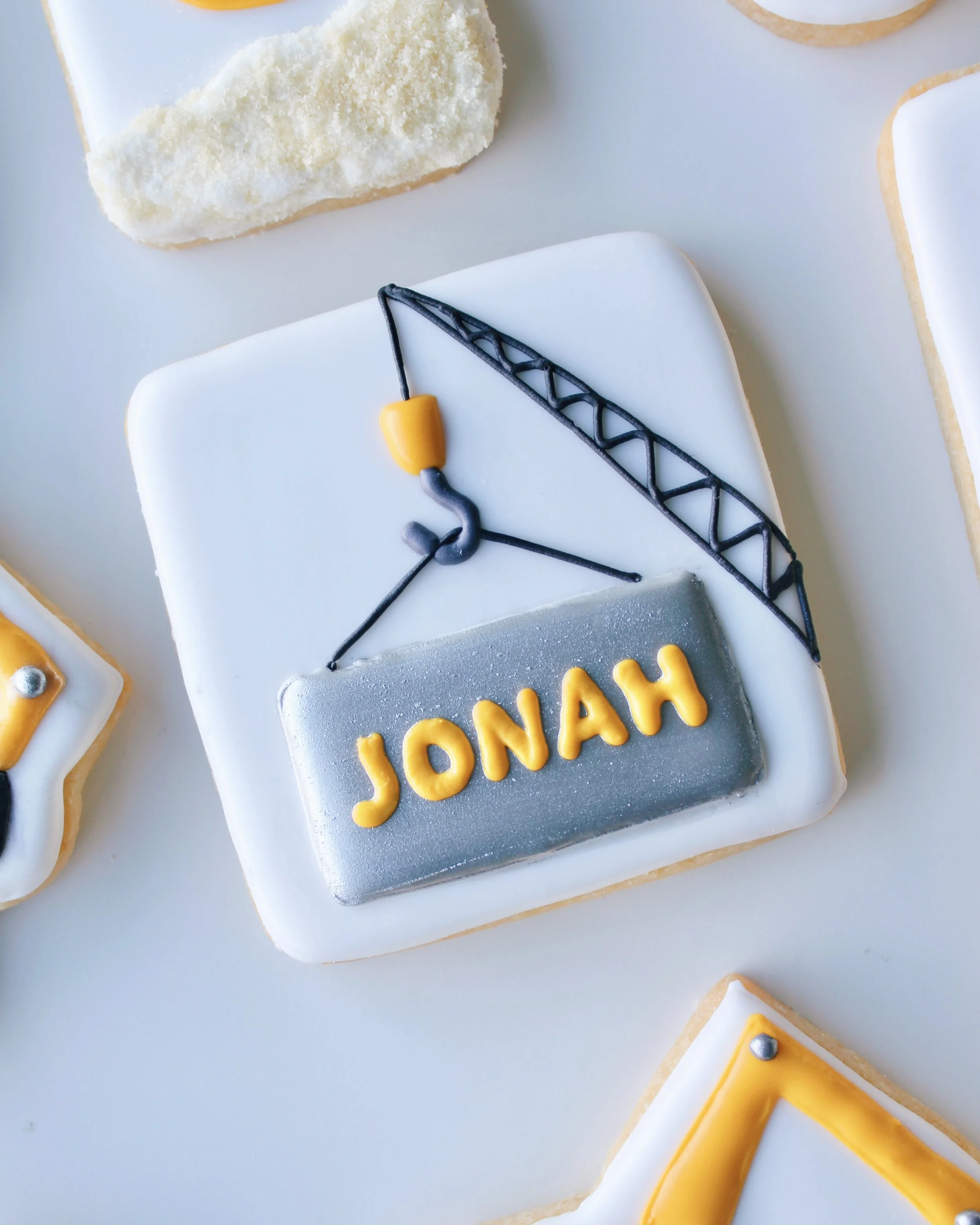 Close-up of silver metal sheet with yellow personalised writing reading “Jonah,” held by a crane arm on a construction birthday cookie featuring trucks, cranes and excavators.