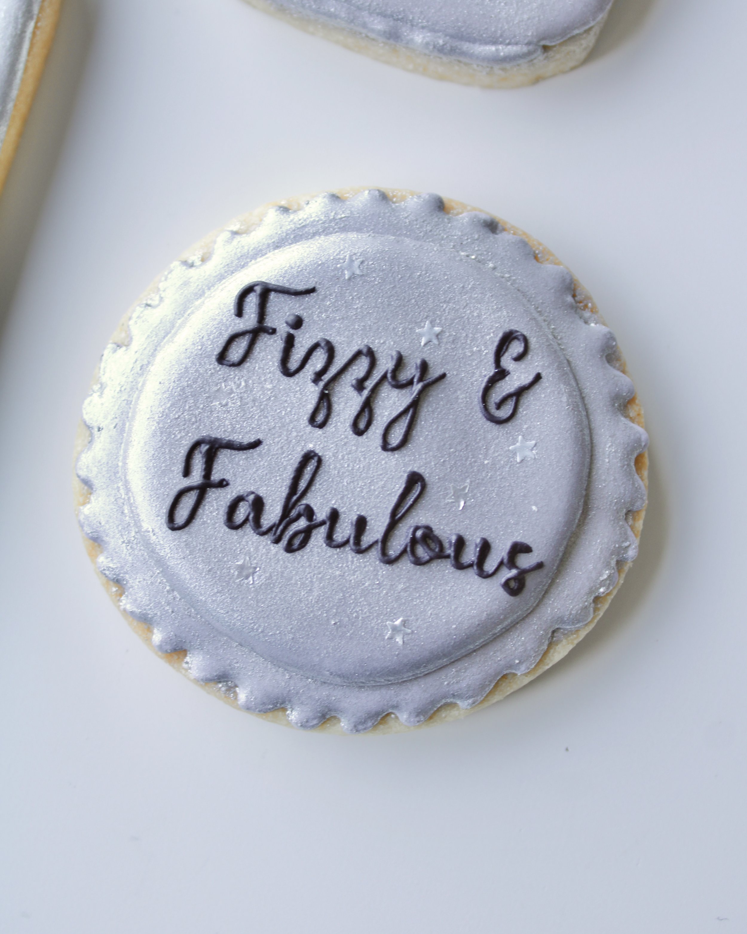 Close-up of a bottle cap cookie with the words “Fizzy and Fabulous” in black script icing, featuring silver star accents.