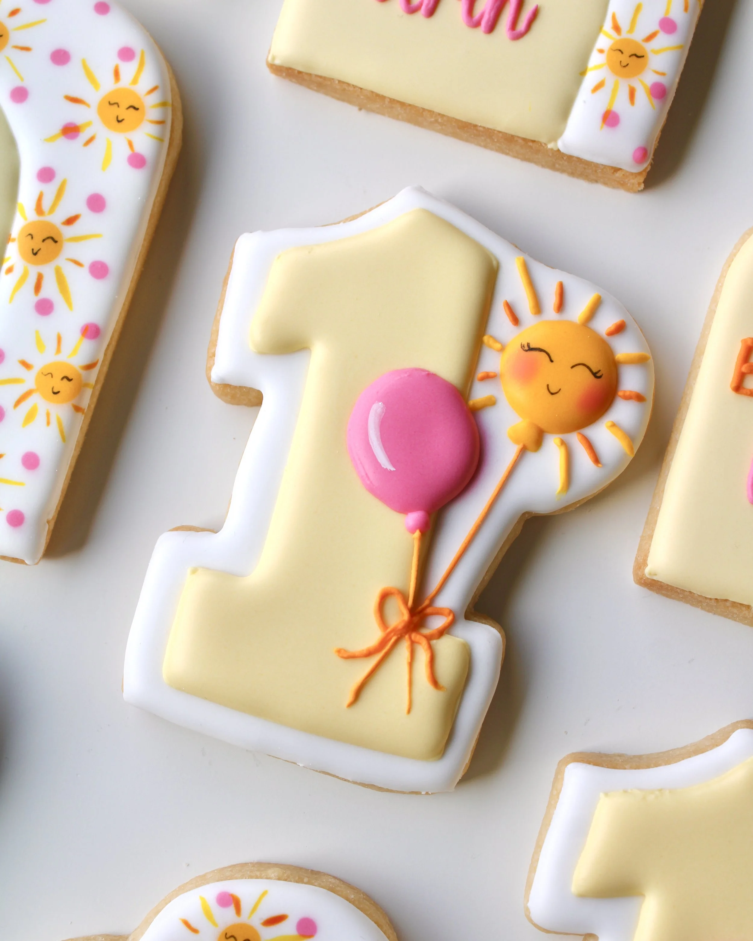 Close-up of a number 1 cookie iced with butter yellow icing, decorated with a pink balloon and a sun-shaped balloon design.