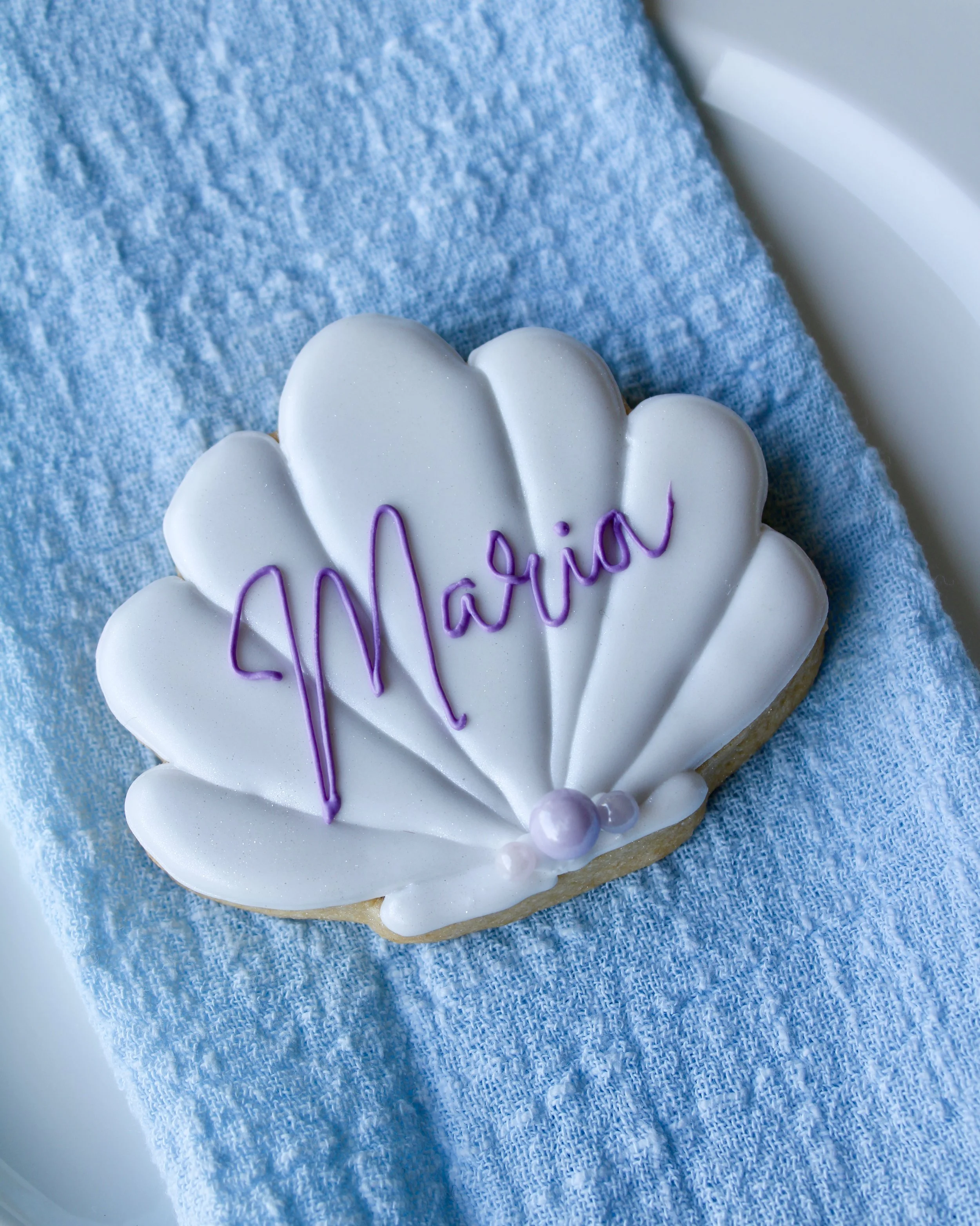 Close up of white seashell cookie with the name “Maria” written in purple cursive icing, placed on a white vintage plate over blue linen napkin and purple crinkled fabric with scattered seashells.