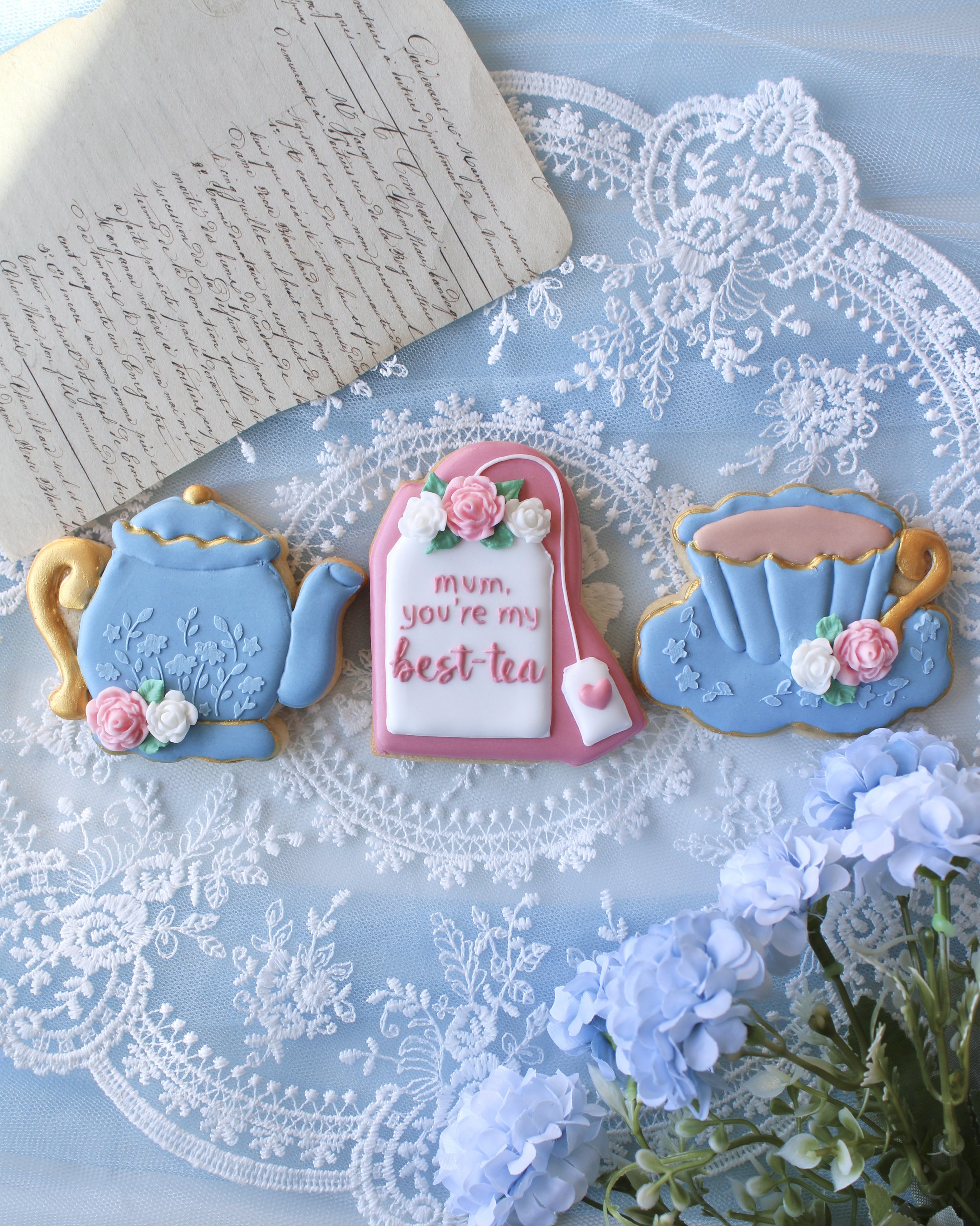 Decorative cookies in the shapes of a teapot, a teabag that reads "mum, you're my best-tea"with flowers, and a cup on a lace tablecloth next to a bouquet of white hydrangeas, with a note on aged paper in the background.
