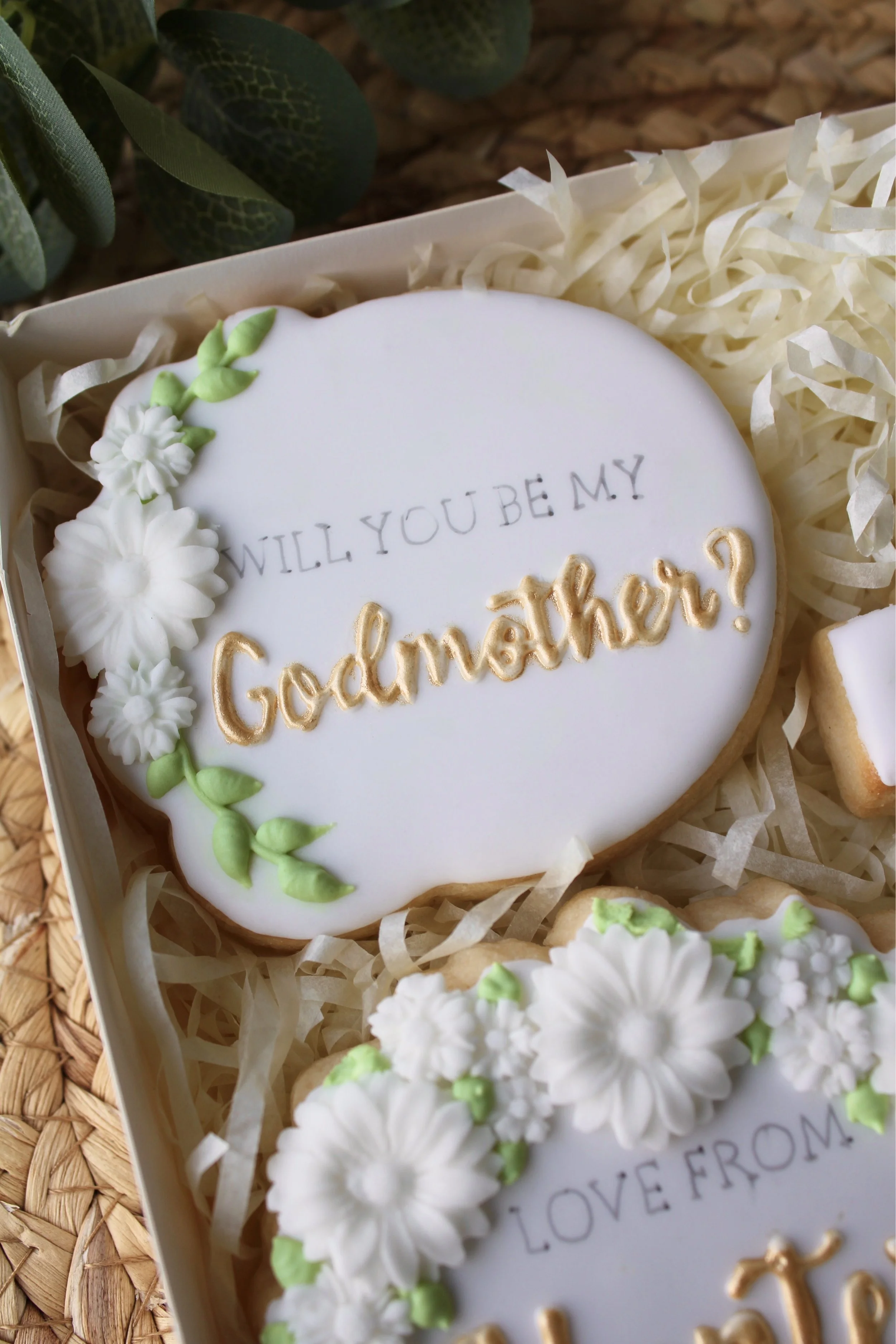 Close-up of a round white iced cookie reading “Will you be my Godmother?” with delicate leaf and white flower decorations.